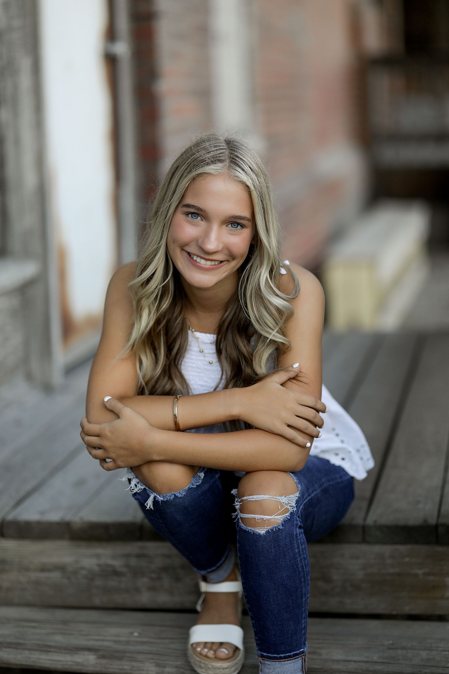Portrait of someone in ripped jeans and white top sitting on wooden steps with a bright genuine smile.