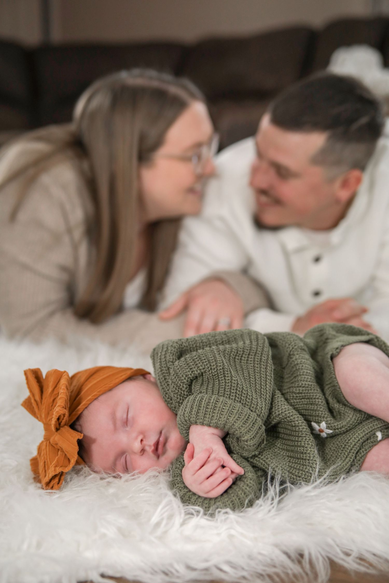 A newborn baby in a green knit sweater sleeps peacefully on a white fur blanket while parents share a tender moment.
