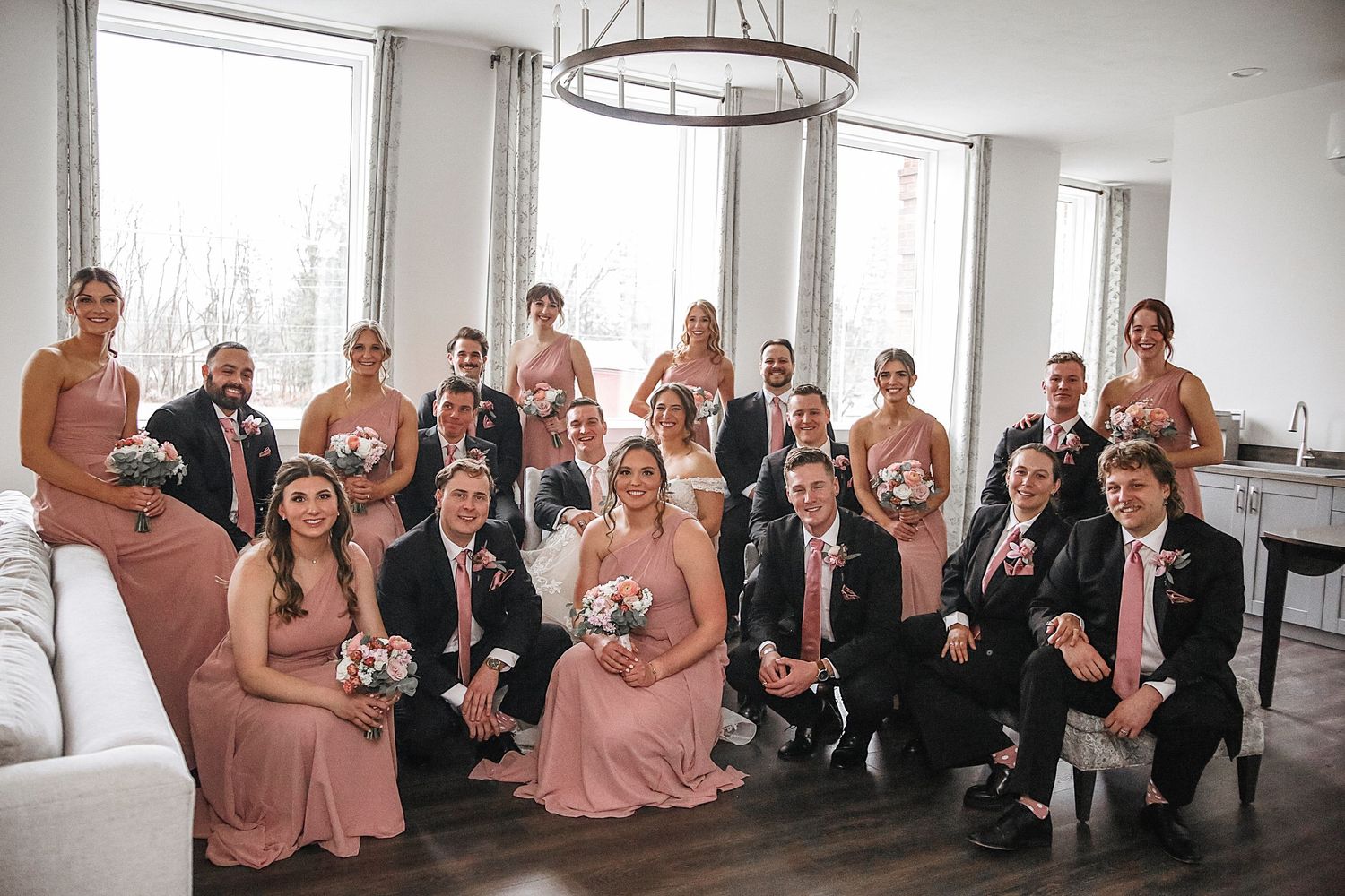 Wedding party poses together in a modern indoor venue with large windows and circular chandelier lighting.
