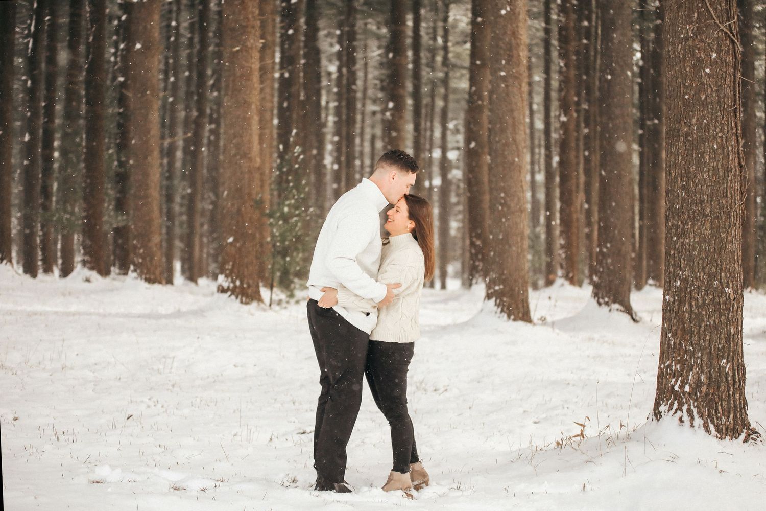 A couple walks together through a snowy forest path with tall pine trees on a winter day.