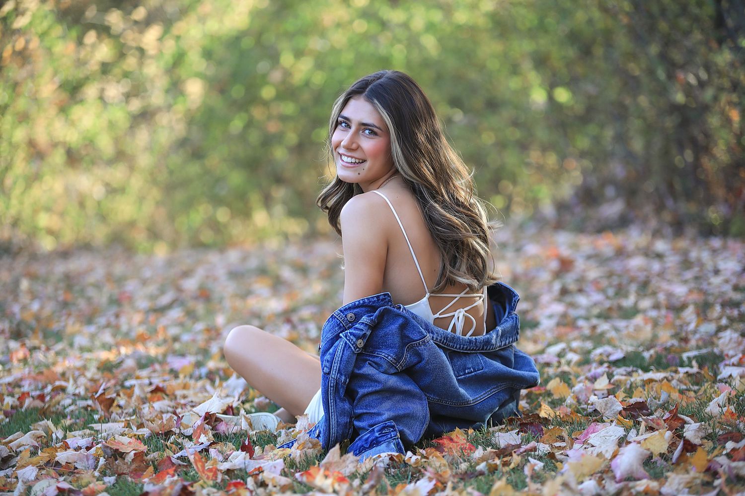 A person lies among colorful autumn leaves wearing a blue dress while smiling on a sunny fall day.
