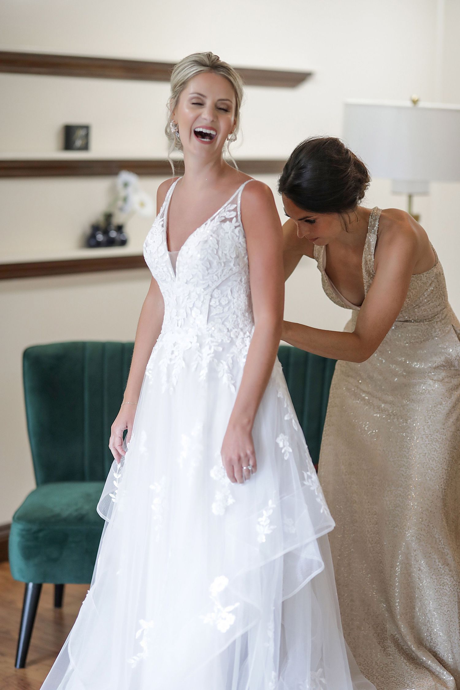 A bride in a white lace wedding dress gets assistance with final adjustments in a room with a green velvet chair.
