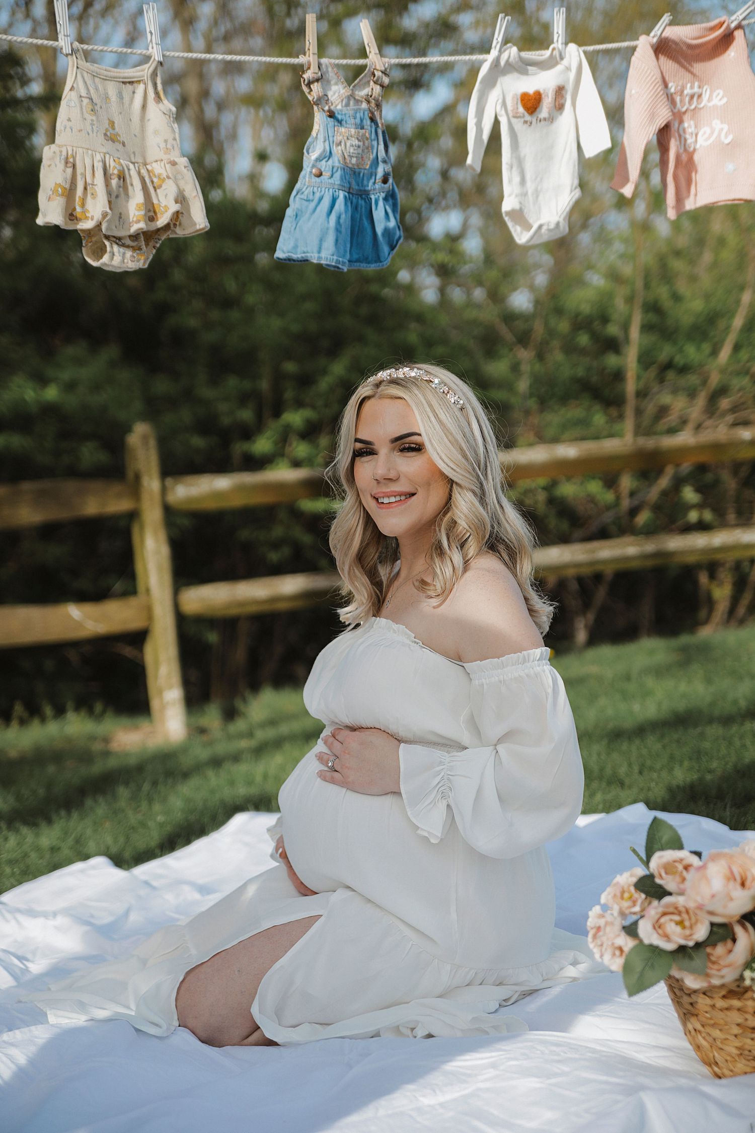 Baby clothes hanging on a clothesline above a blanket with flowers and picnic basket in an outdoor garden setting.