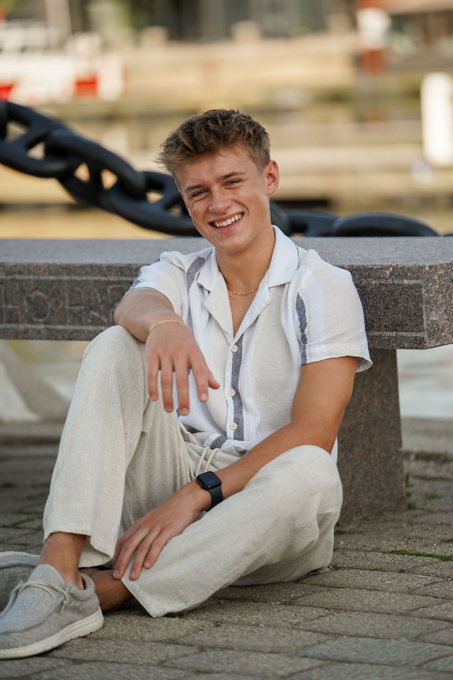 Young person in casual white outfit smiling while sitting outdoors on stone steps near a waterfront.