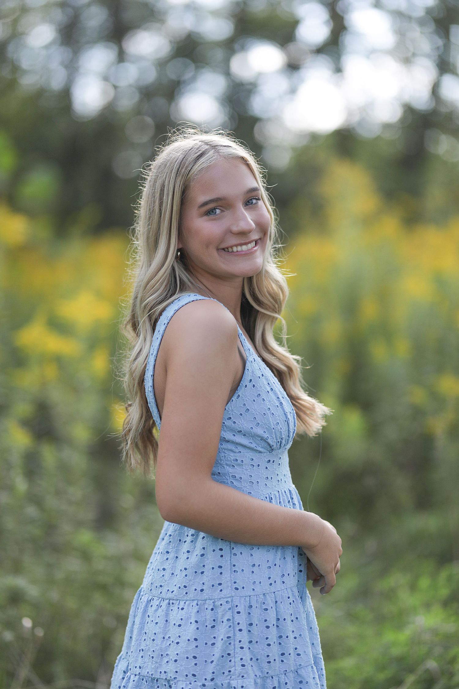 A person wearing a light blue eyelet dress poses outdoors against a blurred nature background with yellow flowers.
