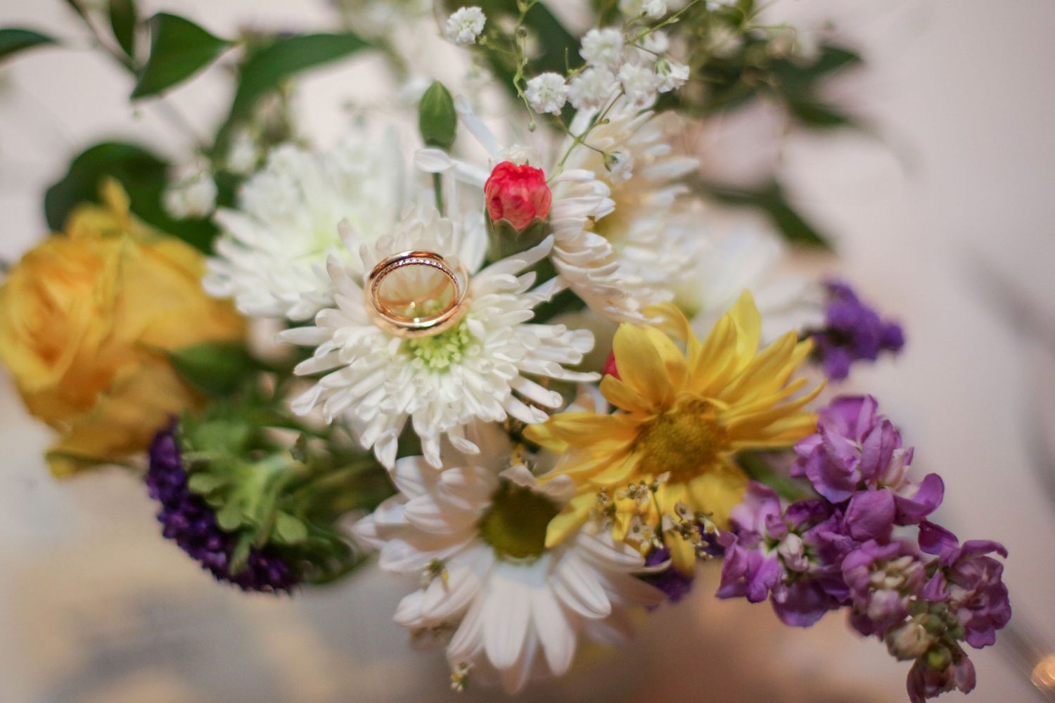Wedding rings nestled among a colorful bouquet of white daisies, yellow roses, and purple flowers in soft focus.