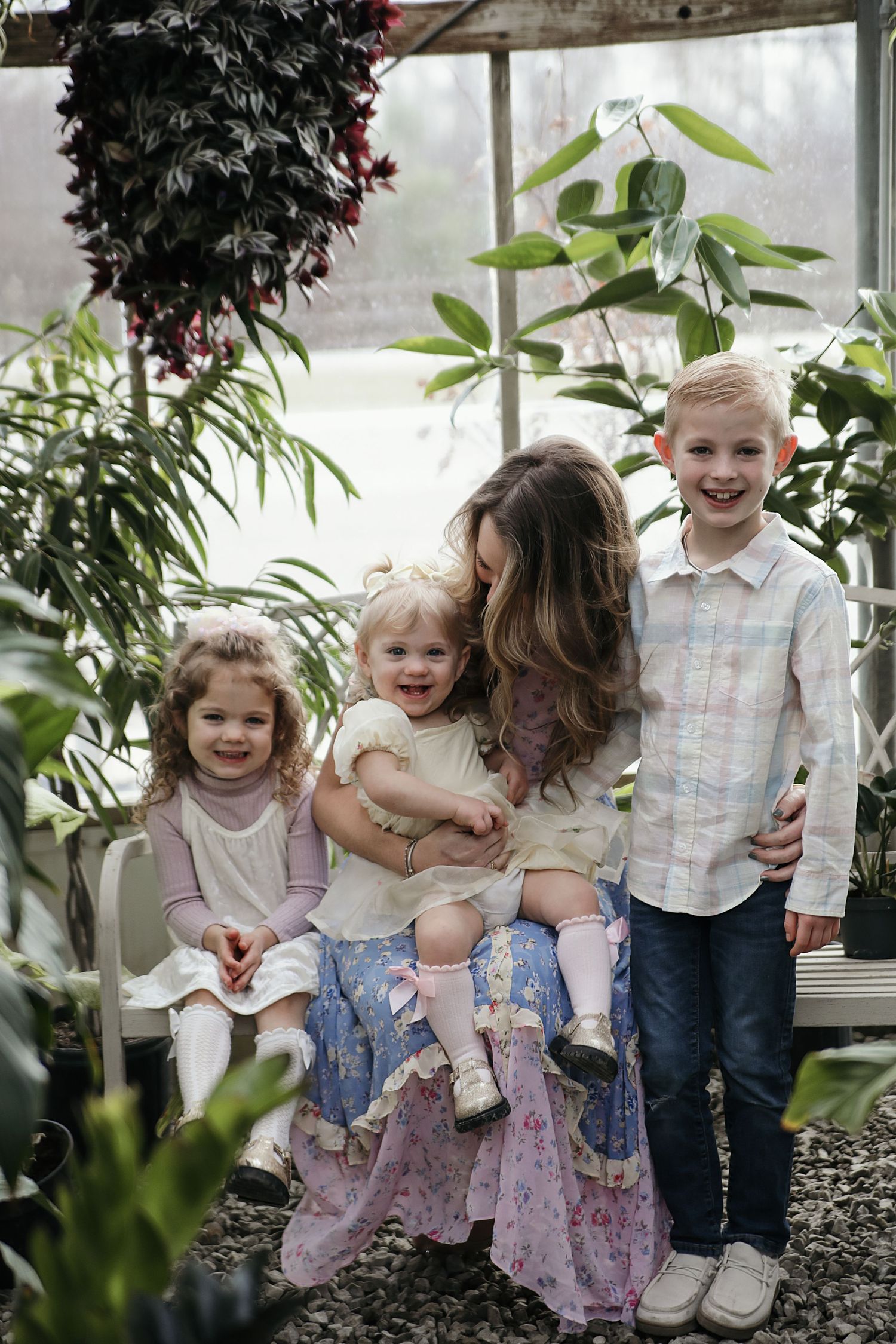 Young children sit together and laugh on a bench in a greenhouse surrounded by lush plants.