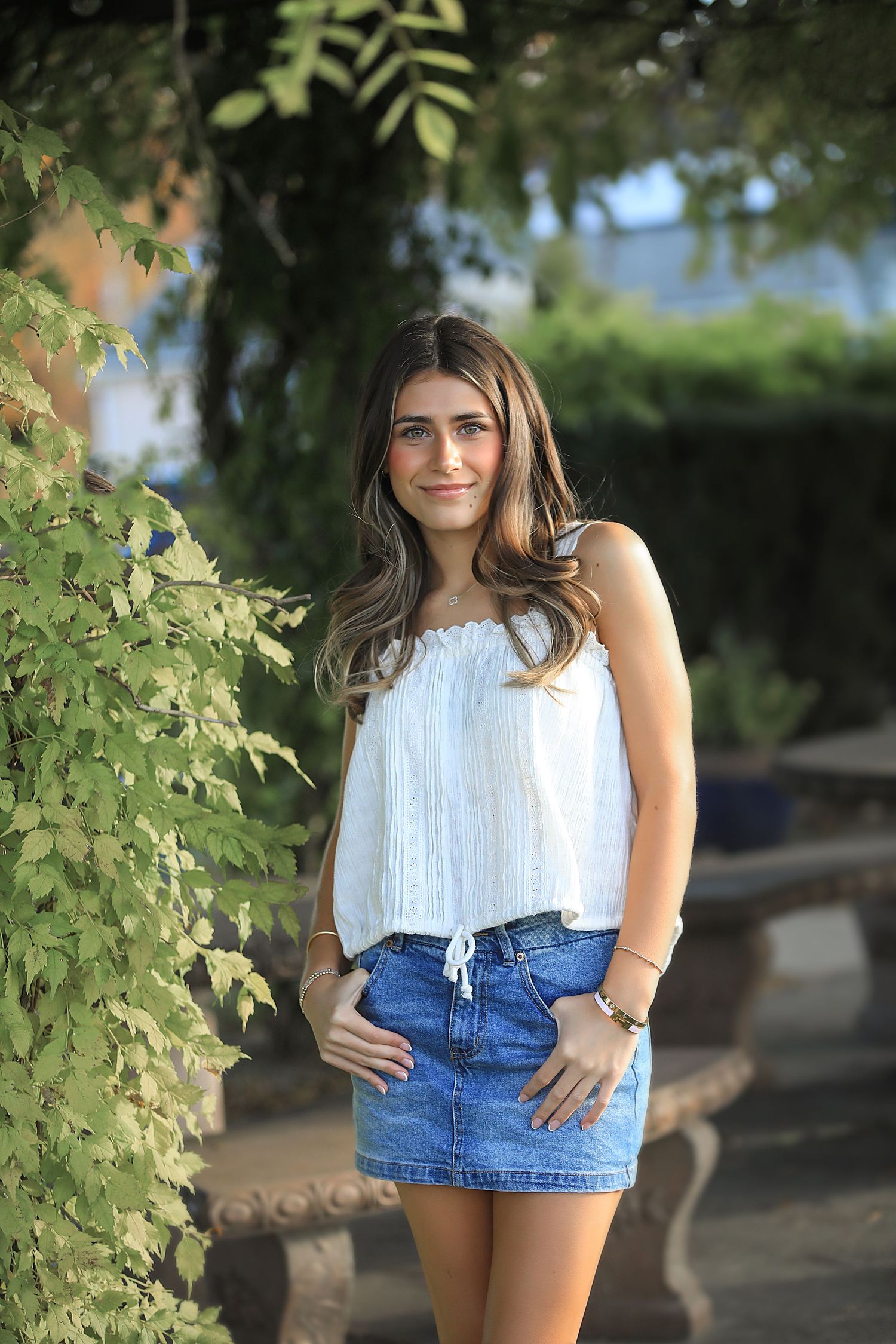 Person in white sleeveless top and denim shorts poses outdoors among green foliage in summer setting.