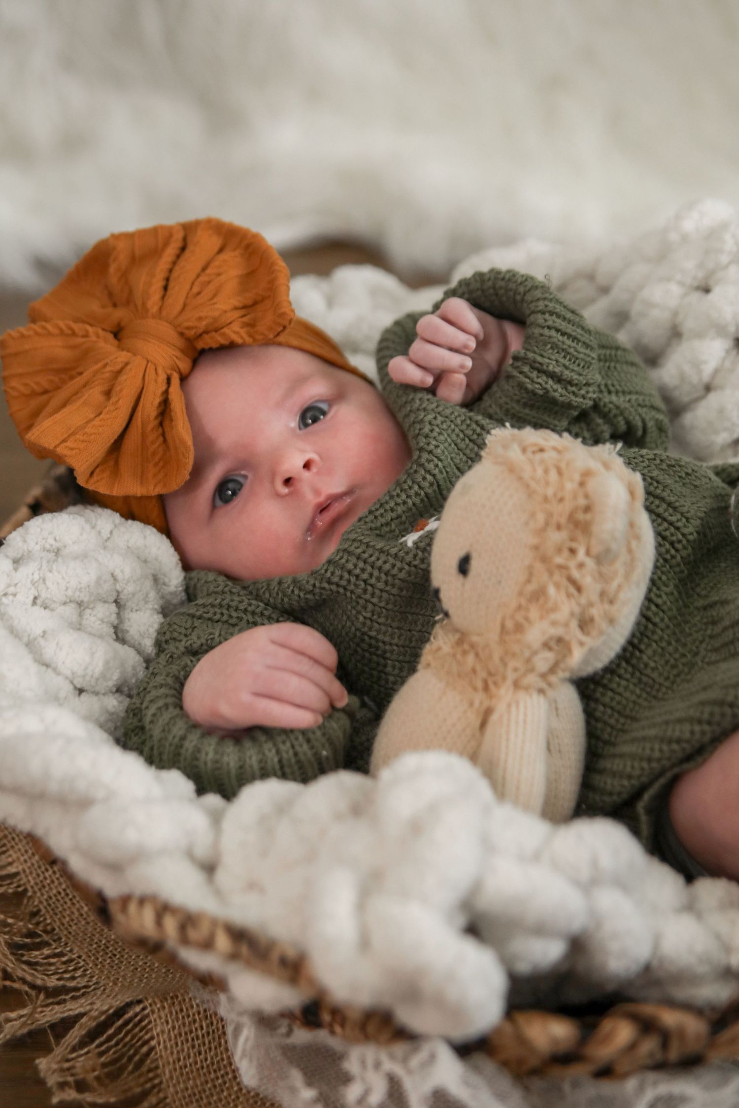 Baby wearing knit green hat and orange bow lies on soft white blanket with plush toy in series of newborn portraits.
