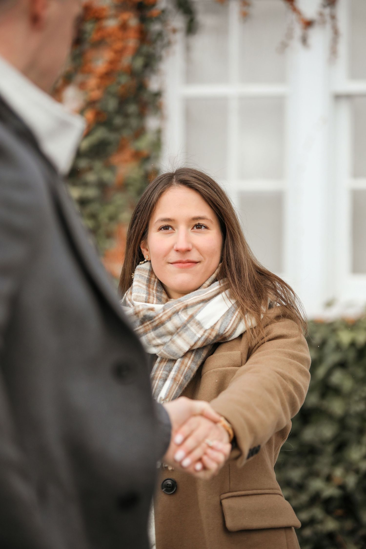 A couple shares a romantic moment outdoors wearing winter coats and plaid scarves against an ivy-covered wall.