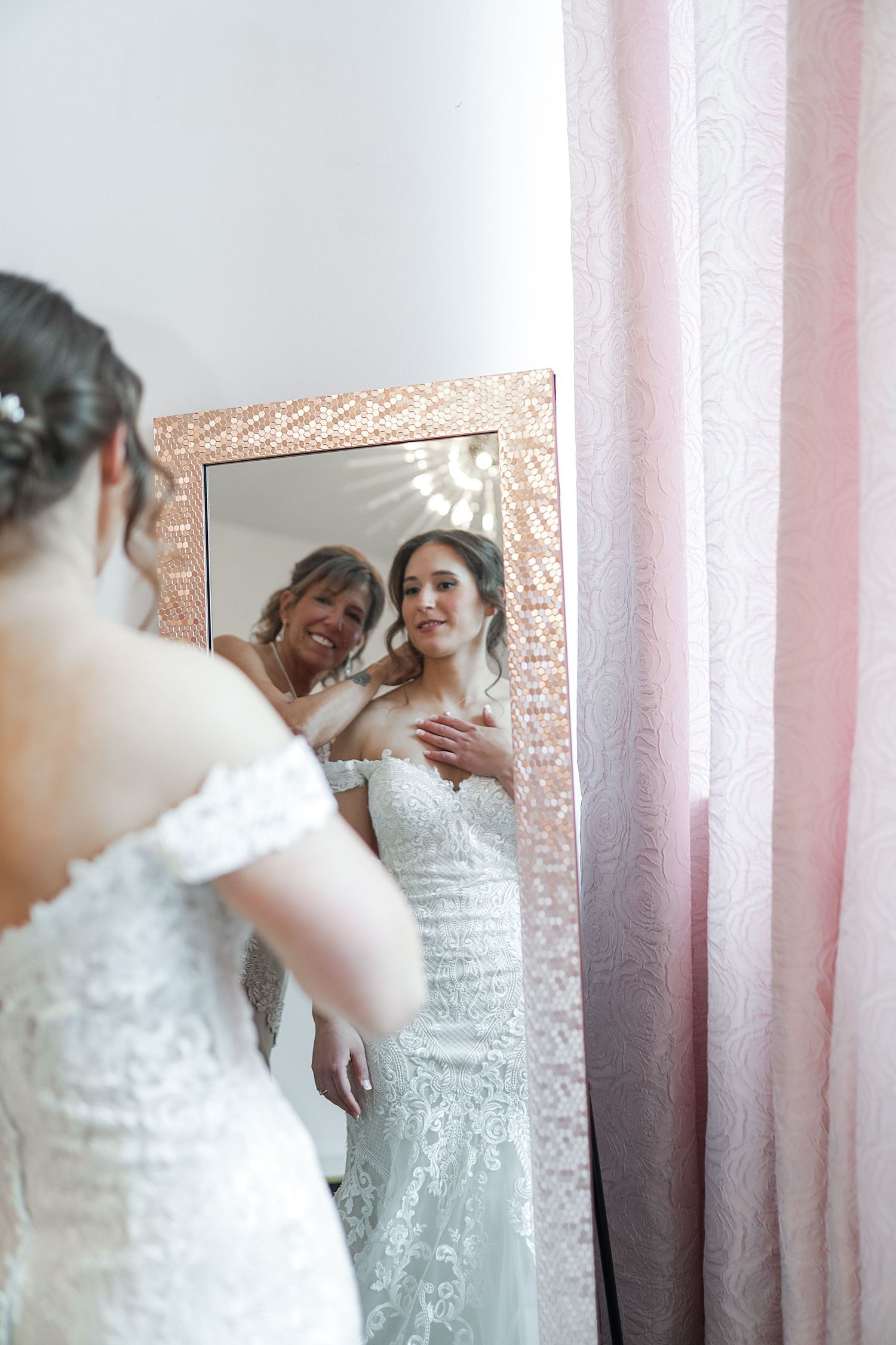 Two people in white dresses share a joyful moment while looking in a mirror with pink curtains in the background.