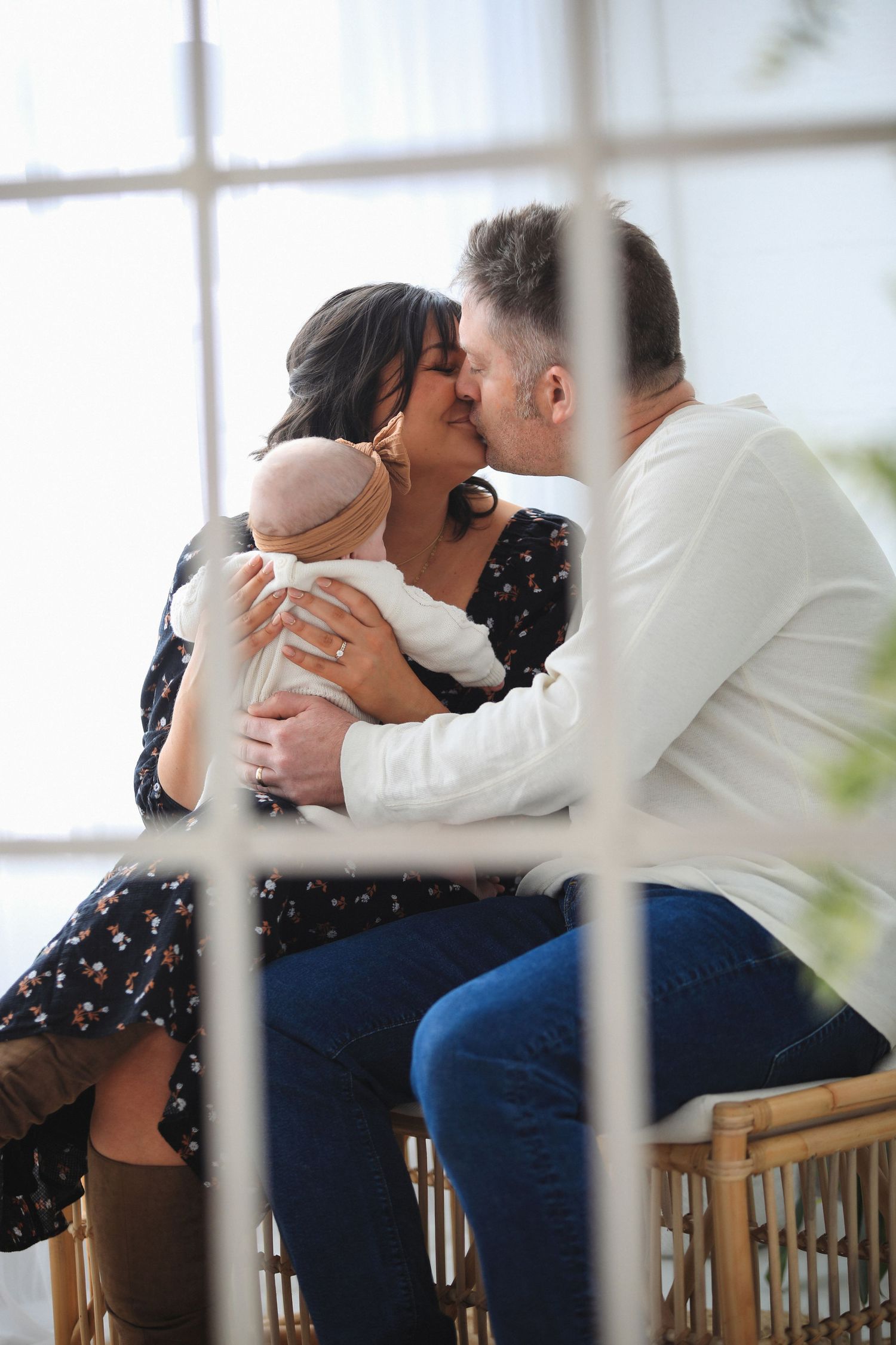 A couple shares an intimate moment while holding a baby in a bright sunlit room near a window.