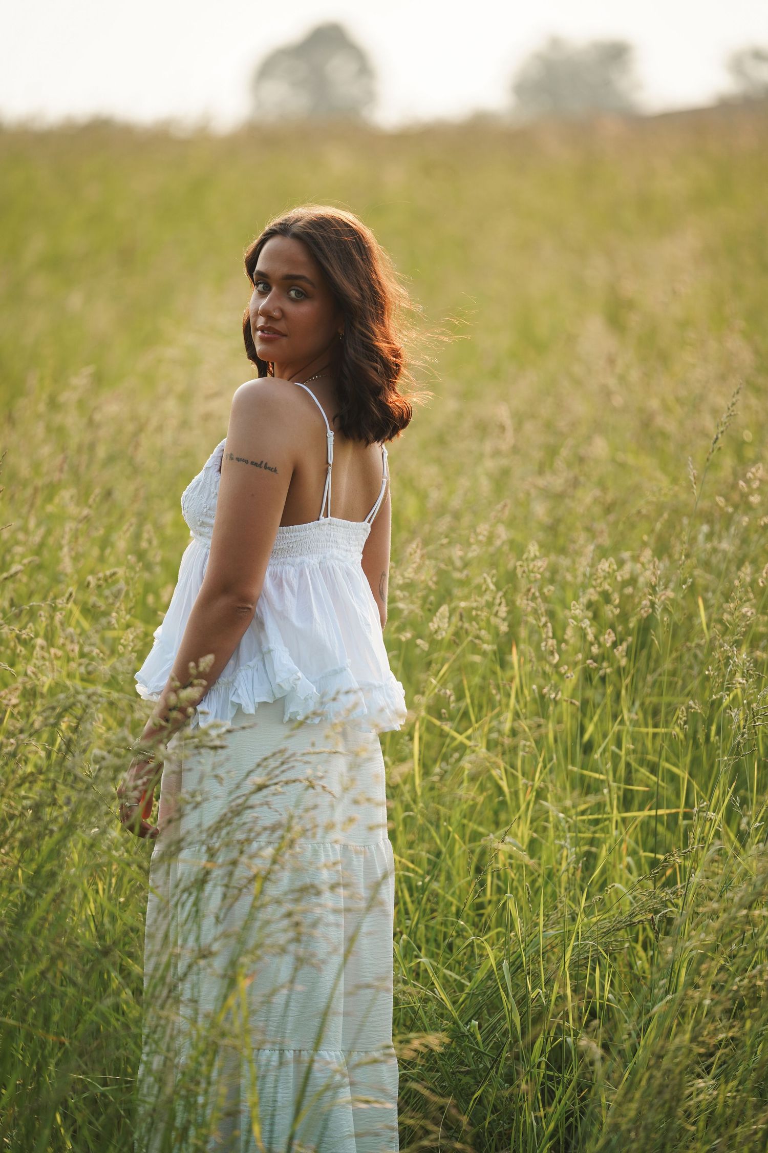 Figure in a flowing white dress poses in a sun-drenched field of tall grass during golden hour.