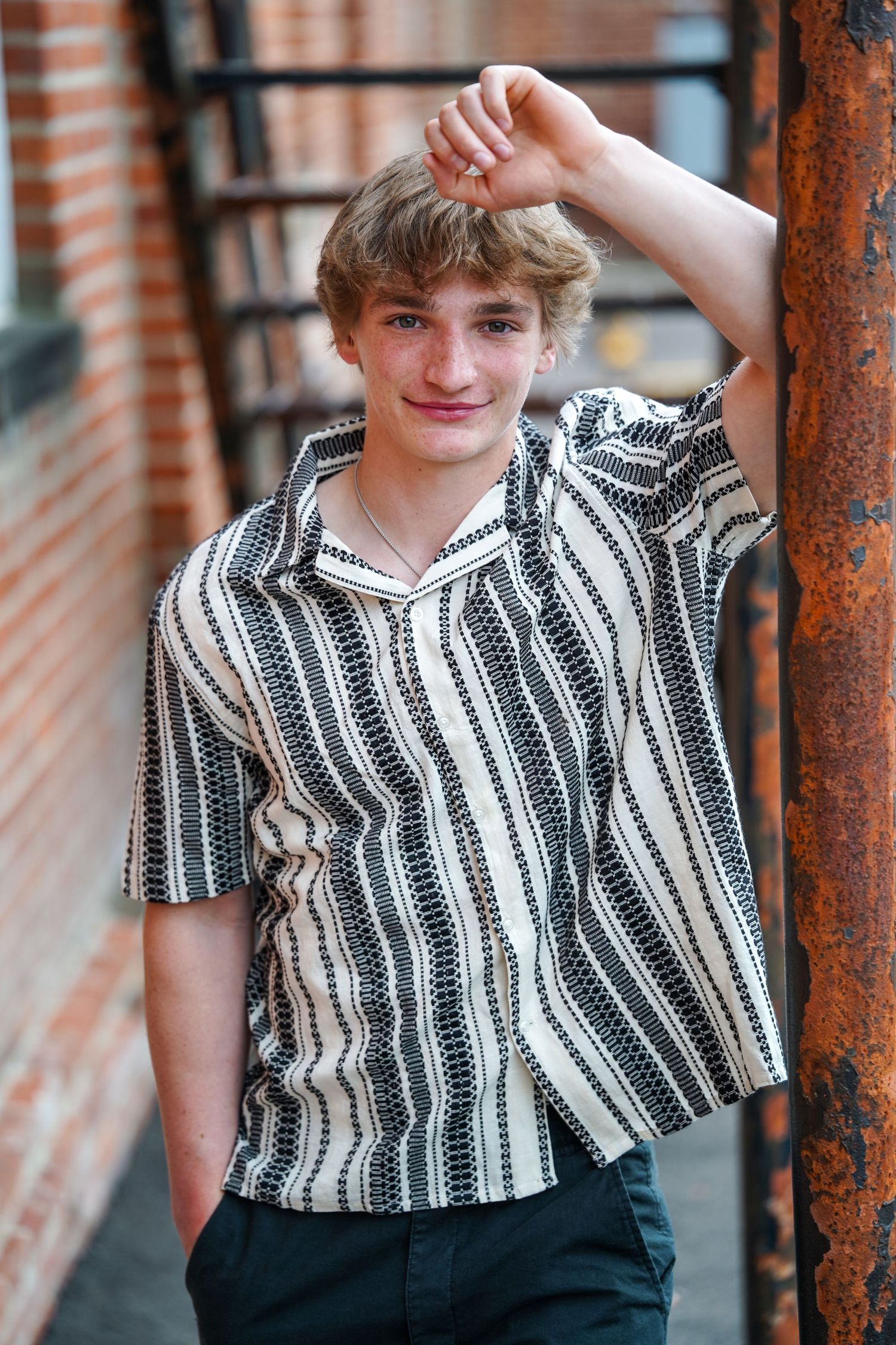 Young student in a black and white striped button-up shirt poses casually against brick wall in outdoor portrait.