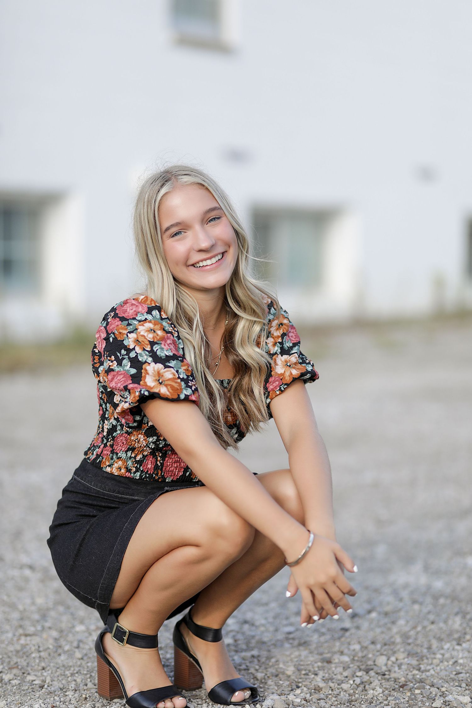 Fashionable woman in floral top and black shorts squats on gravel wearing navy ankle strap heels.