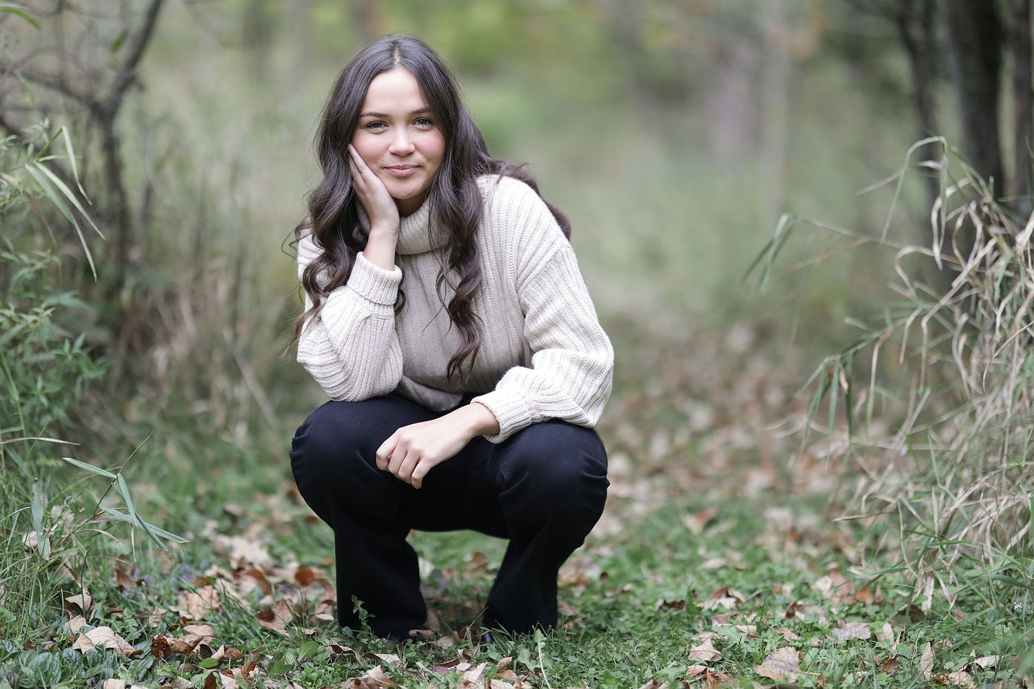 A person in a cream knit sweater and black pants crouches outdoors among autumn leaves and tall grass.