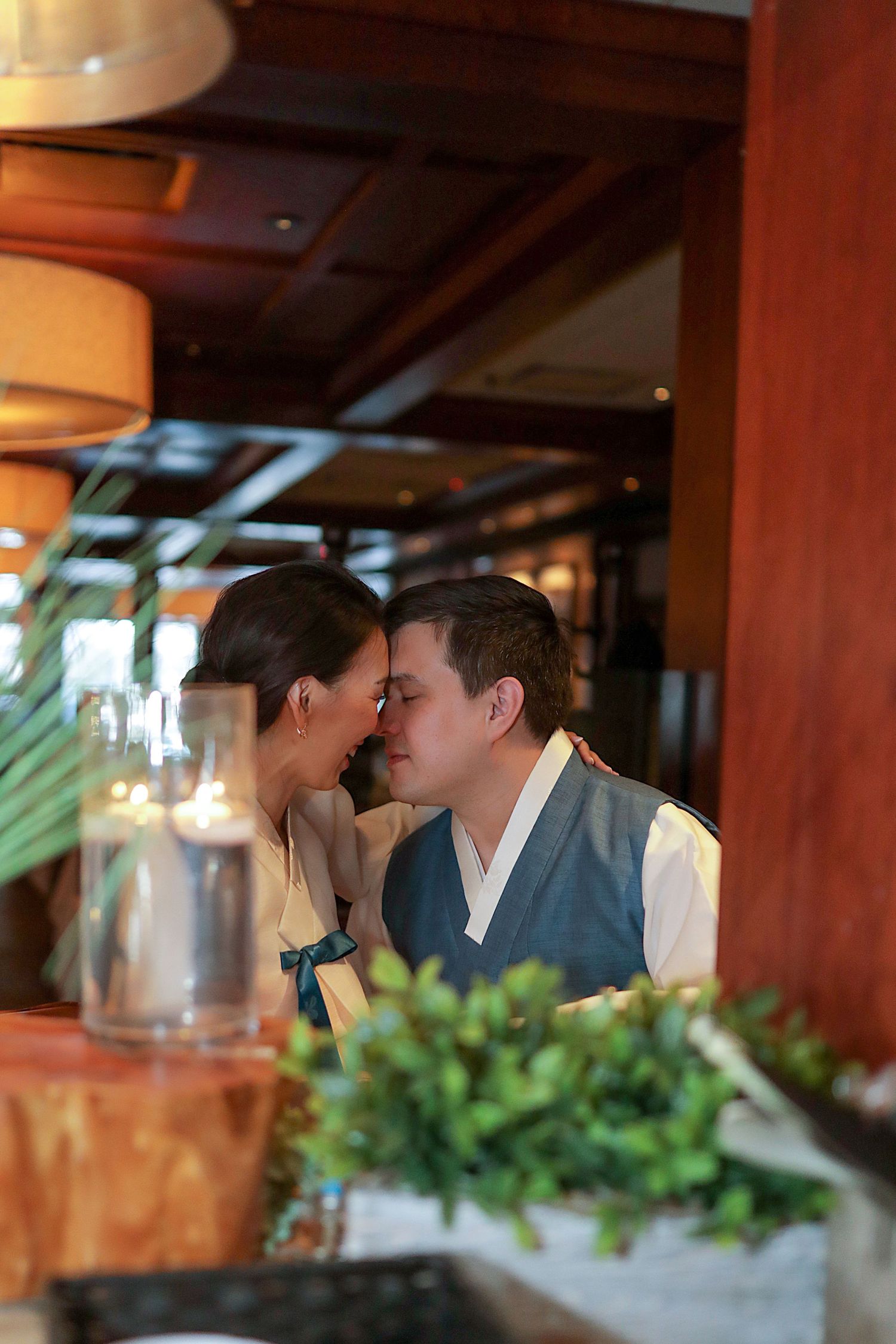 A romantic moment between a couple sharing a tender kiss at a restaurant with warm lighting and greenery in the foreground.
