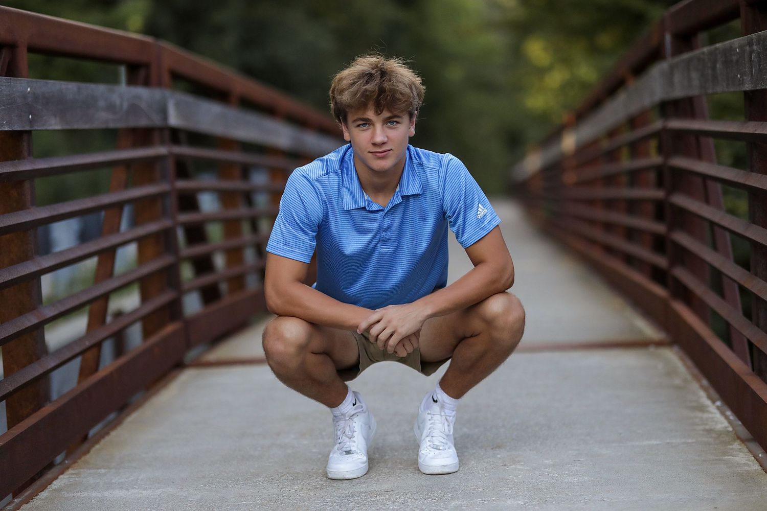 A person in a blue t-shirt and shorts crouches on a wooden bridge in a park setting.