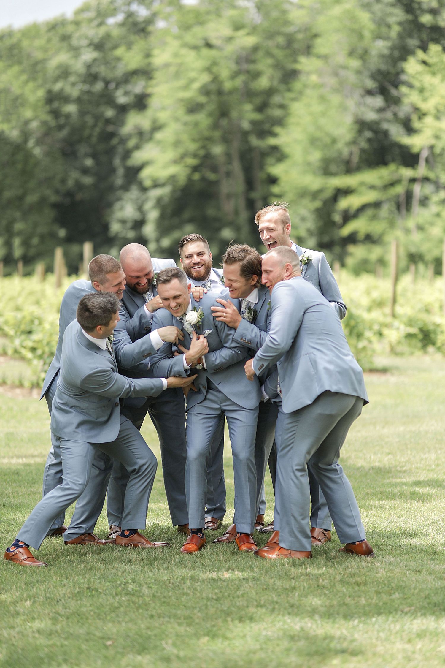 Groomsmen in grey suits pose playfully on grass in a football huddle formation.