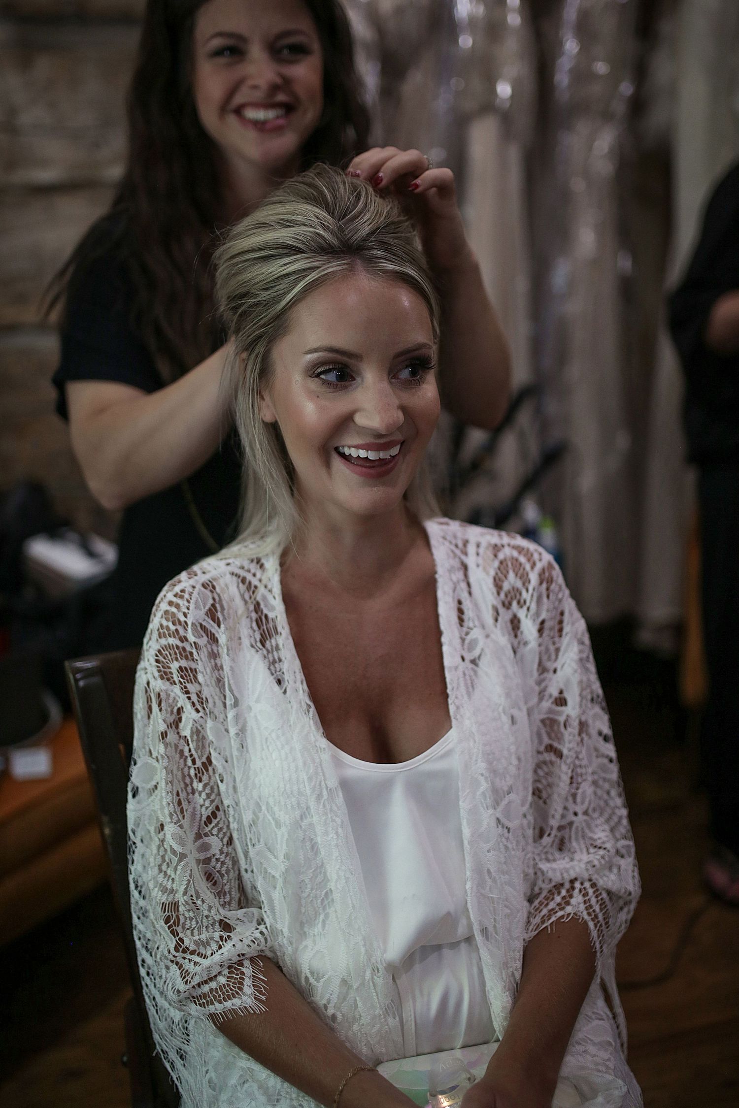 Wedding preparation scene showing a stylist working on bridal hair in a rustic indoor setting.