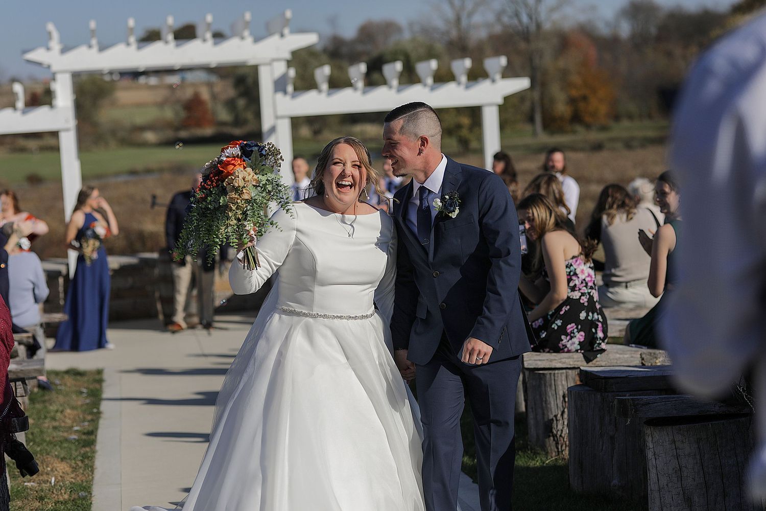 Newlyweds walk under a white pergola during their outdoor autumn ceremony.