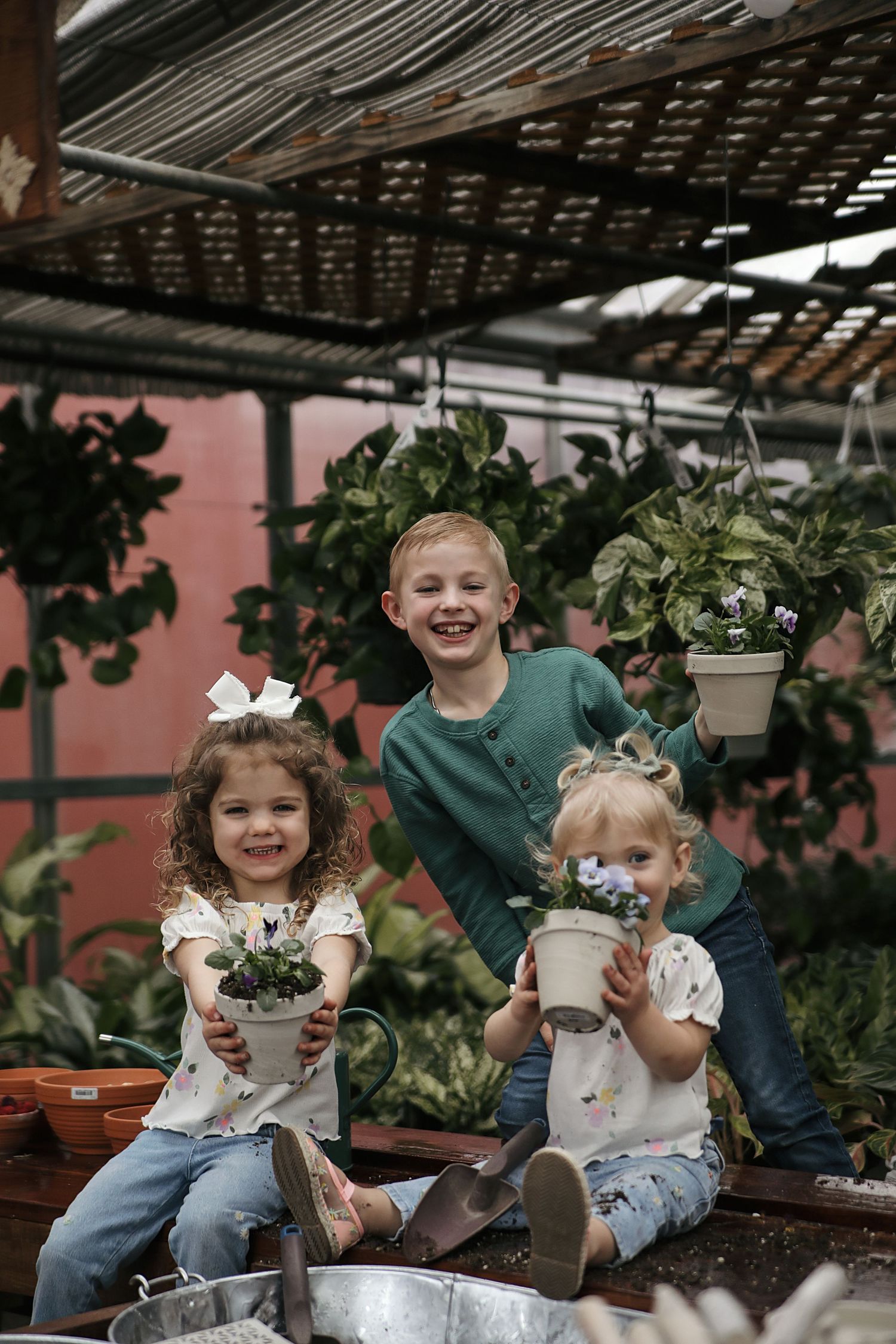 Three children sit in a greenhouse surrounded by potted plants and hanging baskets in a rustic garden setting.