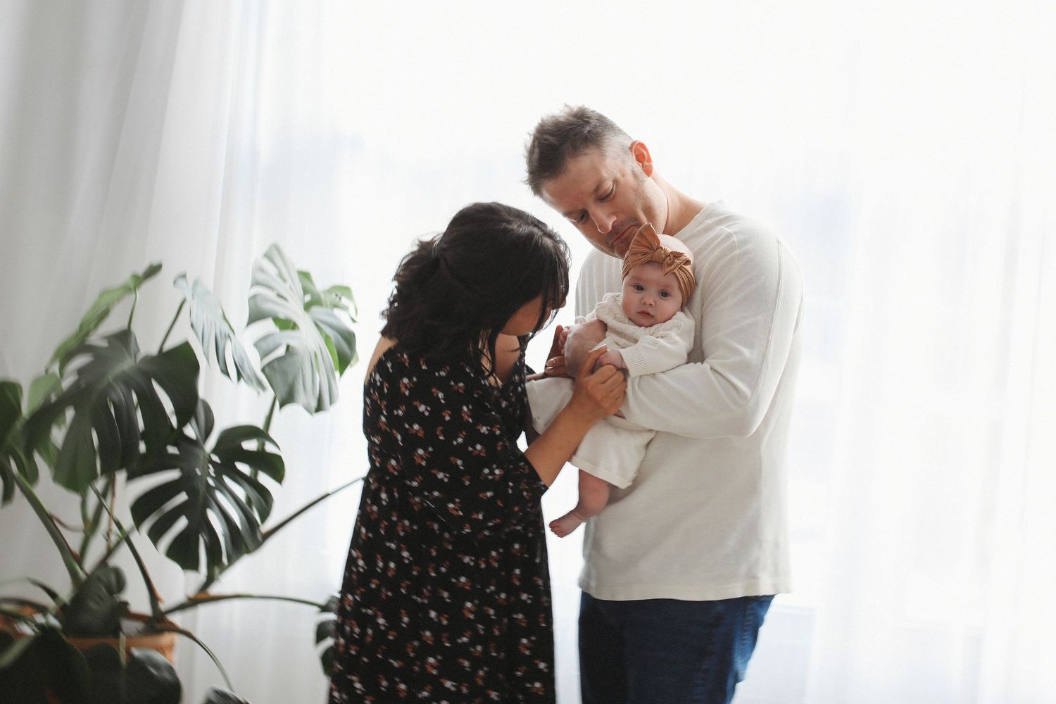 Two adults share a tender moment with a baby in a bright room with monstera plants by the window.