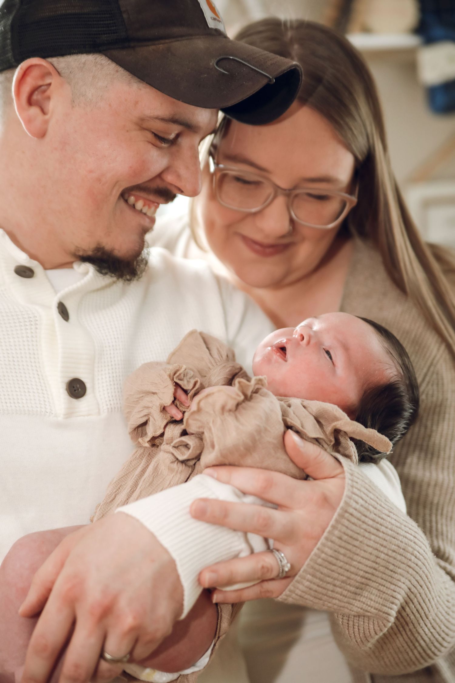 A loving couple smiles while holding their newborn baby wrapped in a beige blanket.