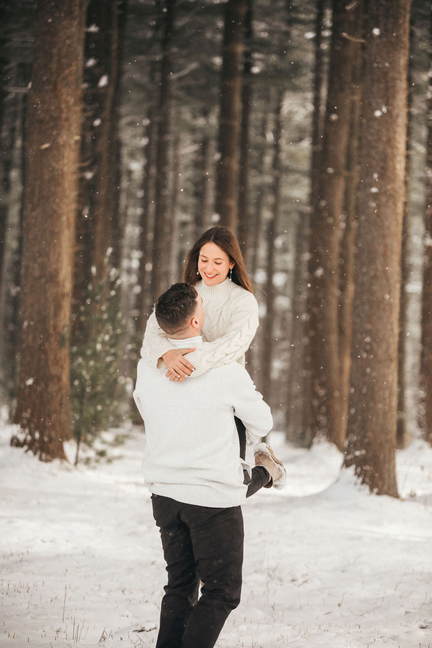 A couple playfully runs through a snowy forest while wearing white and black winter outfits.