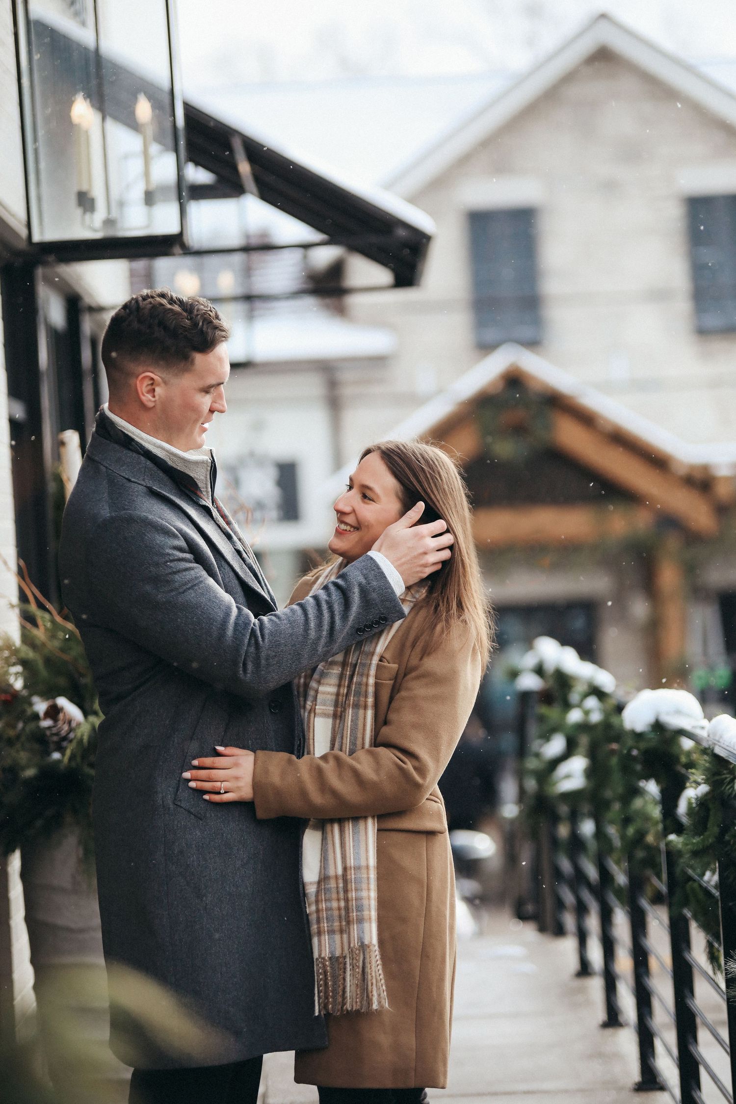 A couple in winter coats share an intimate moment outdoors against a snowy backdrop with houses in the background.
