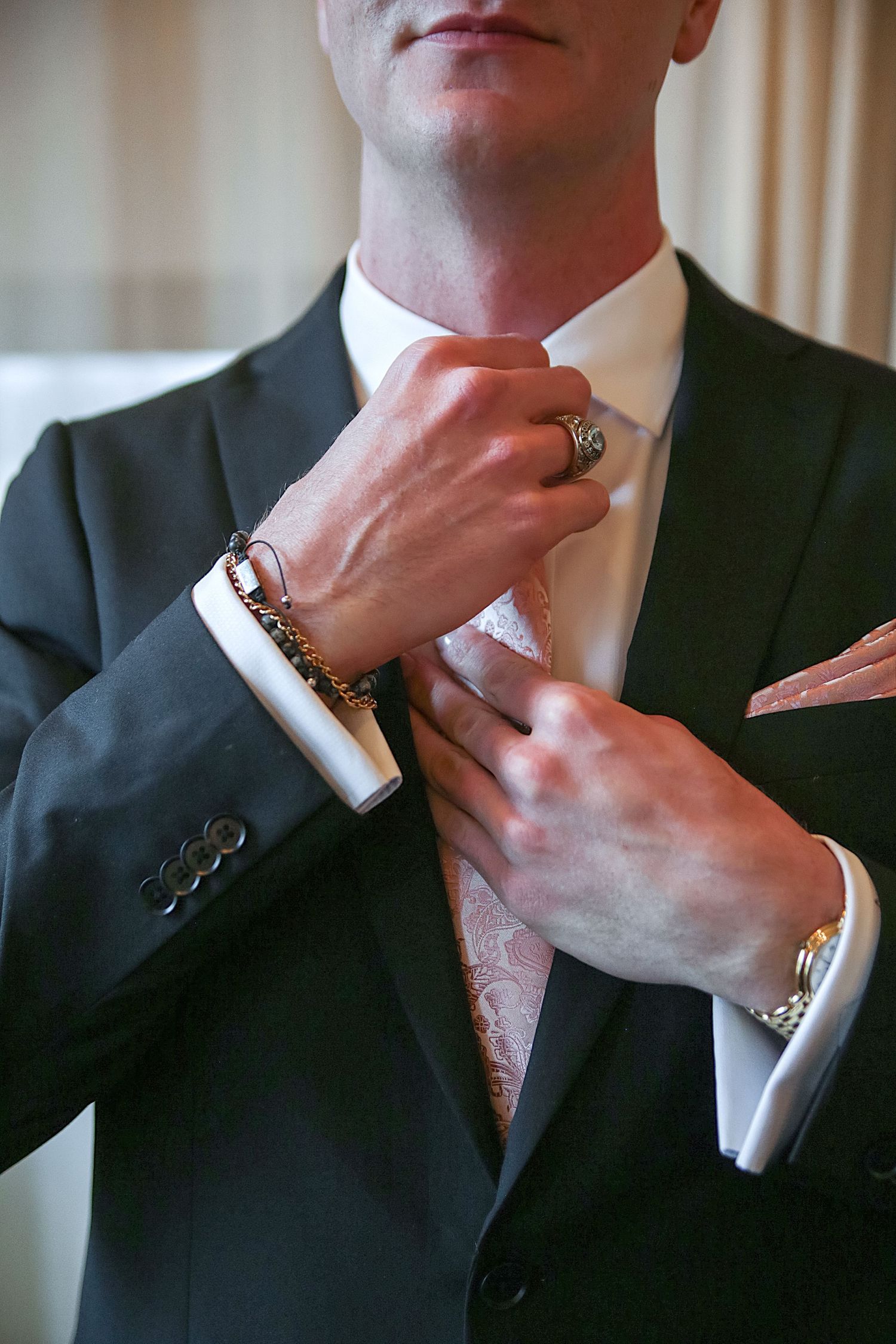 Close-up shot of hands adjusting a tie and suit jacket with jewelry and accessories visible.
