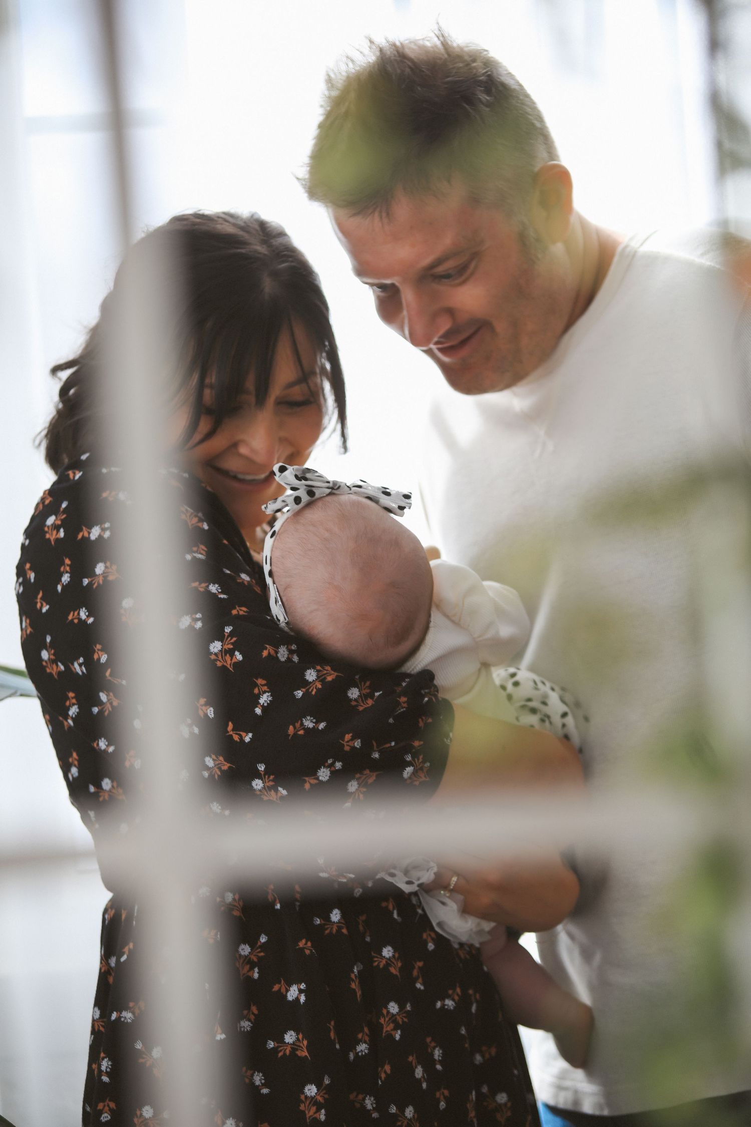 Family shares tender moment with baby in sunlit room with white curtains and greenery.