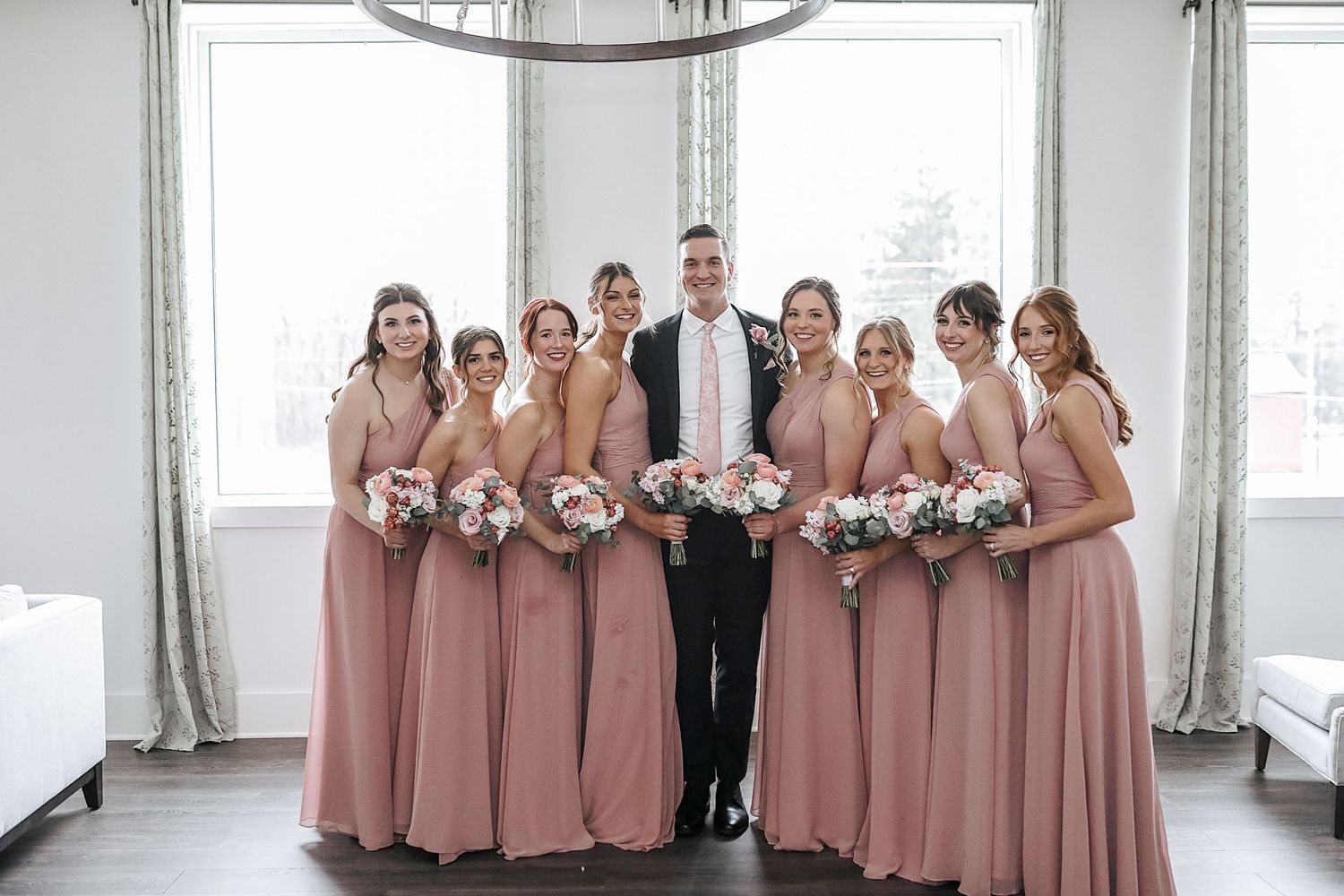 Wedding party in matching dusty rose dresses poses together in a bright indoor venue with large windows.