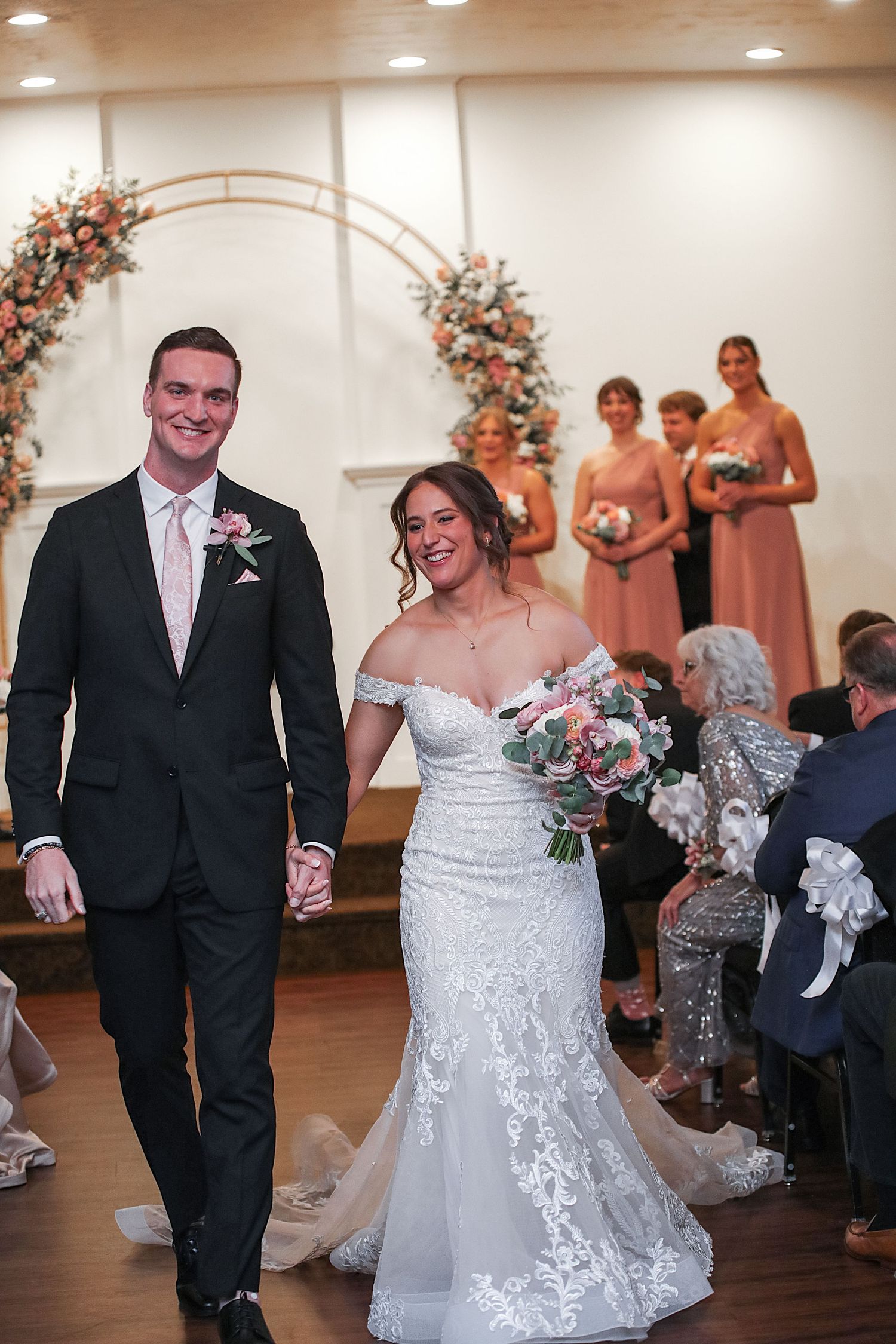 A wedding ceremony with a floral arch backdrop and bridesmaids in peach dresses standing at an indoor venue.