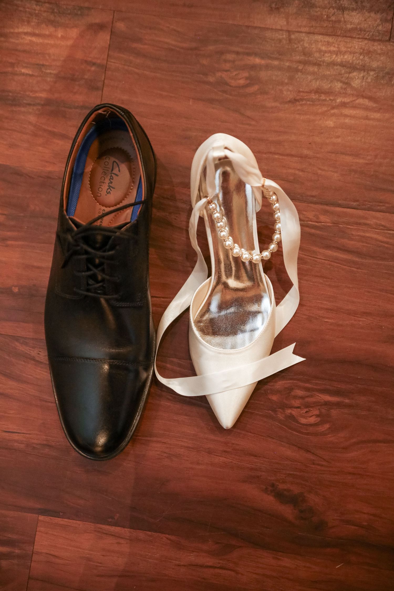 A black leather oxford shoe and white satin bridal heel sit together on a dark wooden floor.