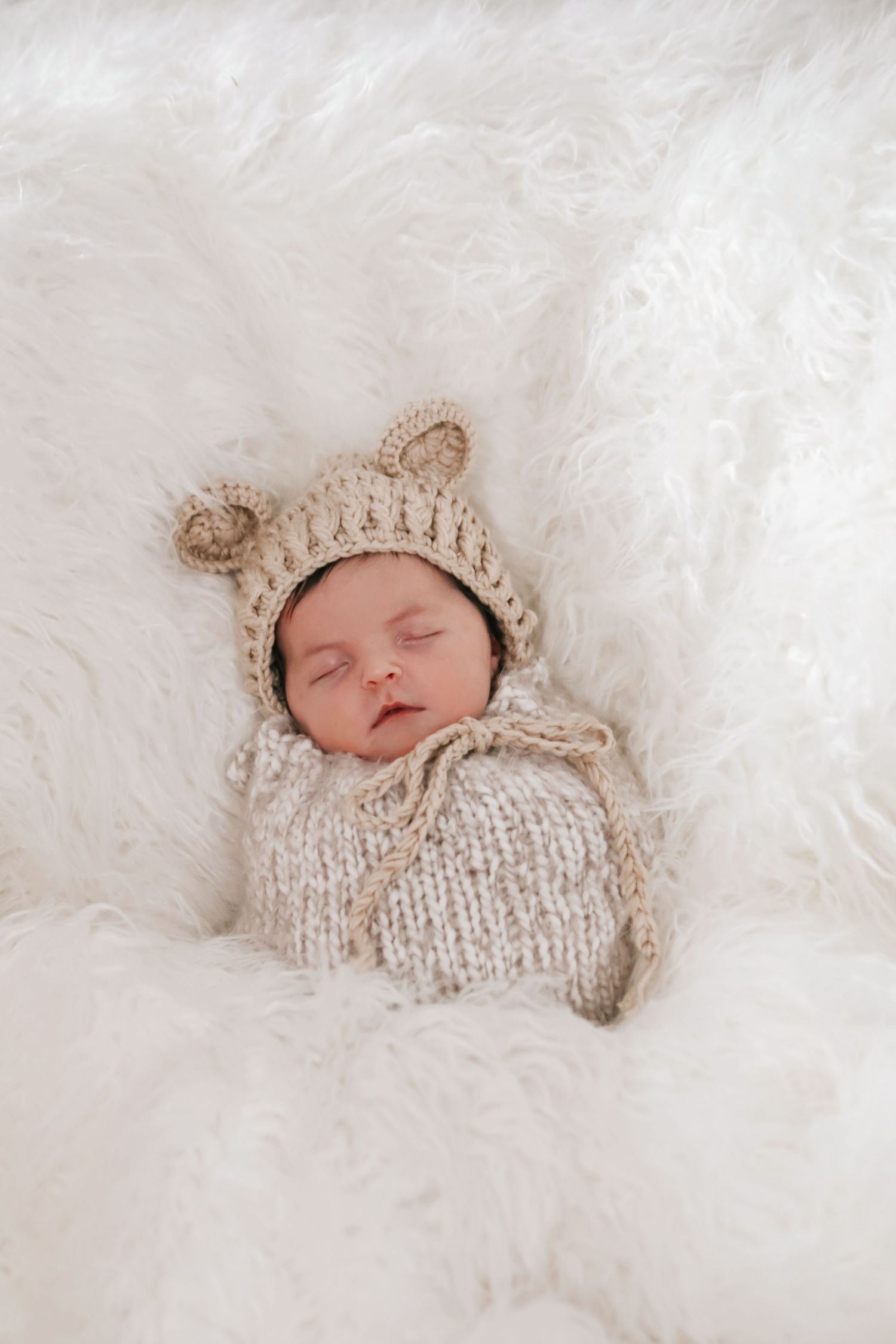 Sleeping newborn wearing cozy knit bear hat nestled in soft white blankets.