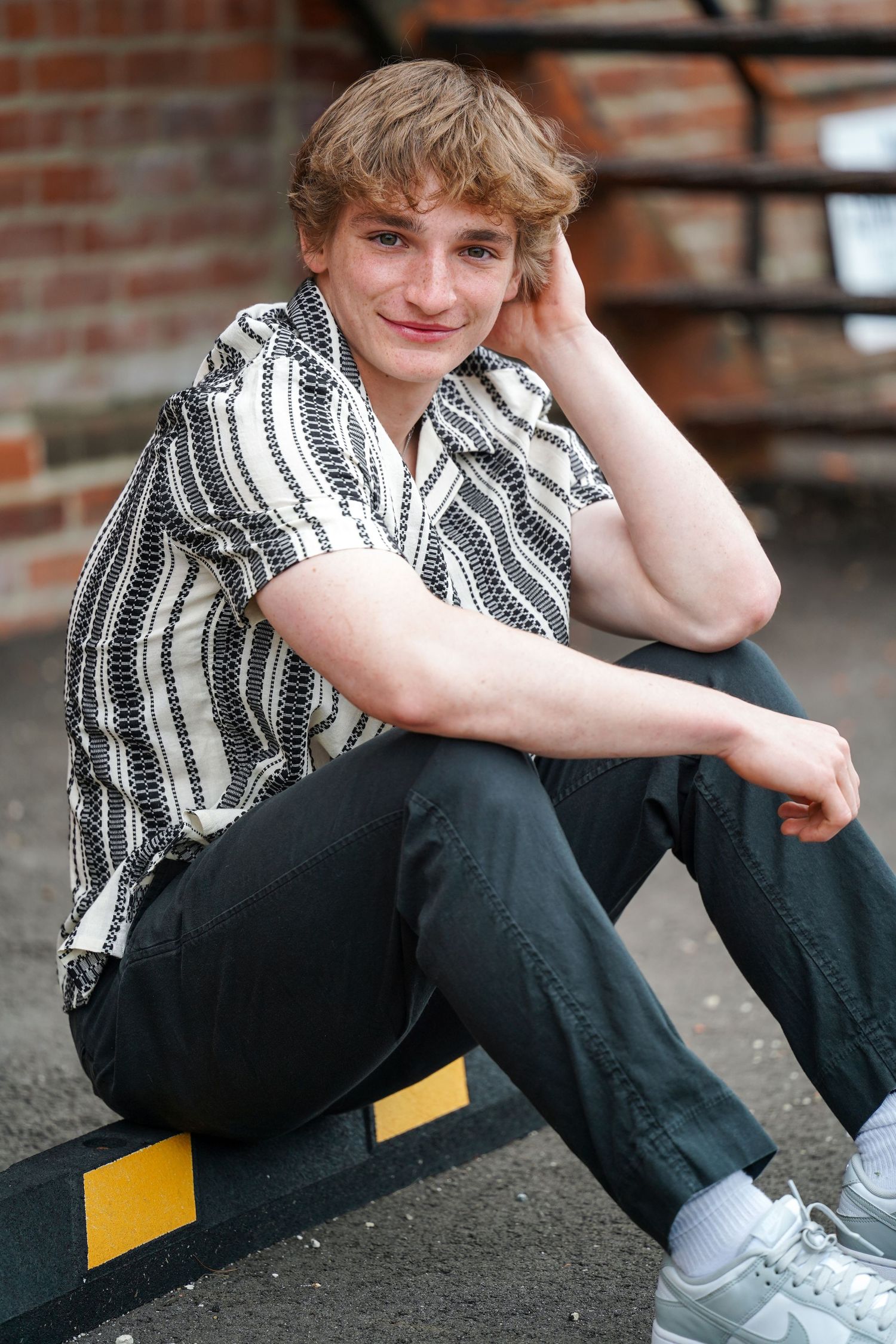 A person in a striped shirt and black pants sits casually against a brick wall wearing gray sneakers.