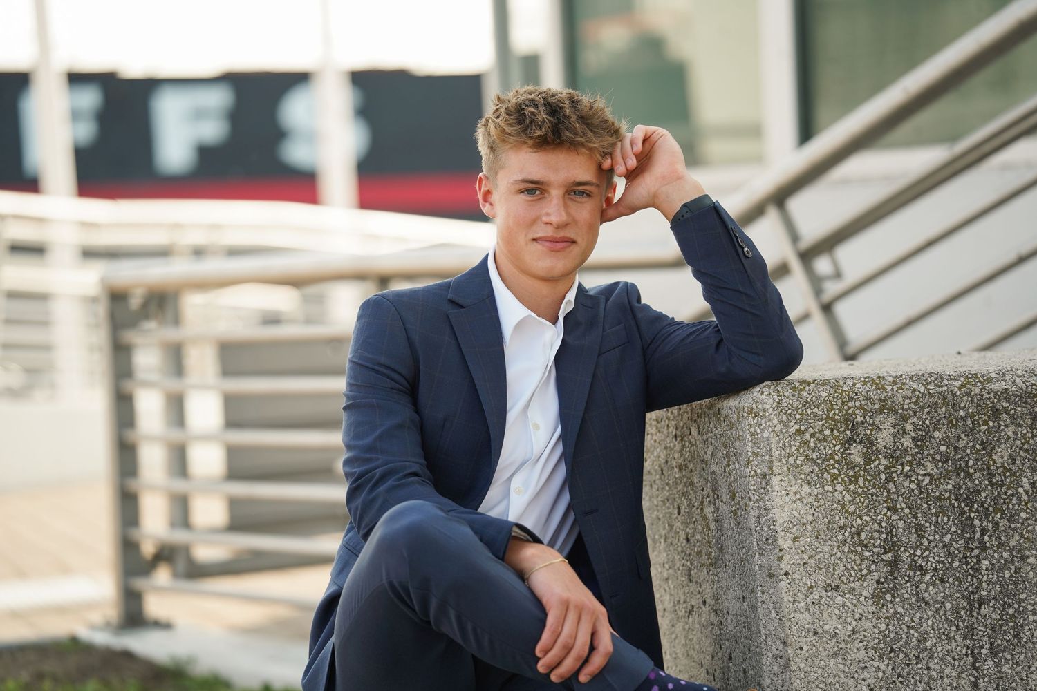 A person in a navy suit and white shirt sits casually on concrete steps while touching their hair outdoors.