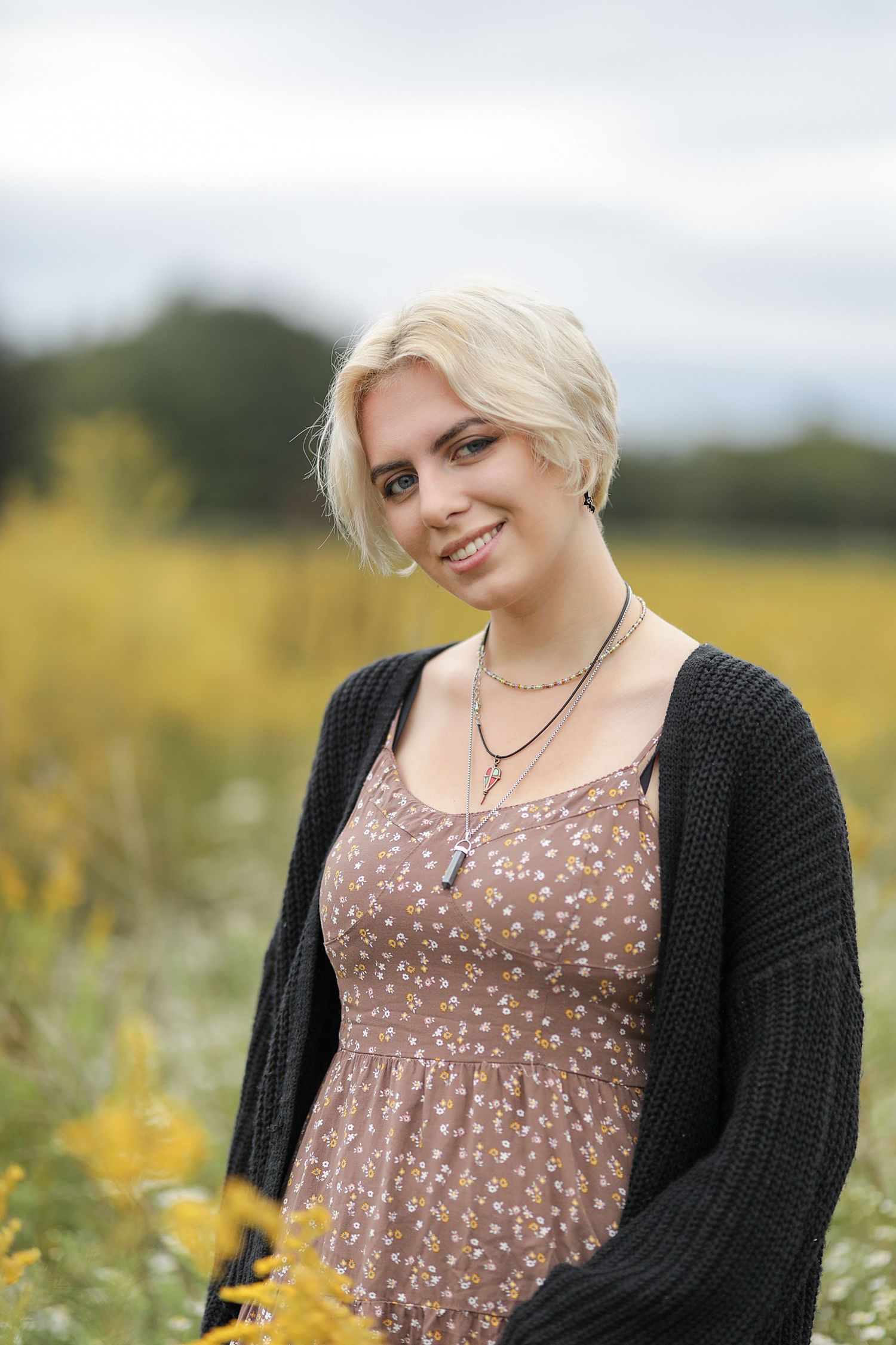 Elegant portrait in a wildflower field with short blonde hair, floral dress and black cardigan on an overcast day.