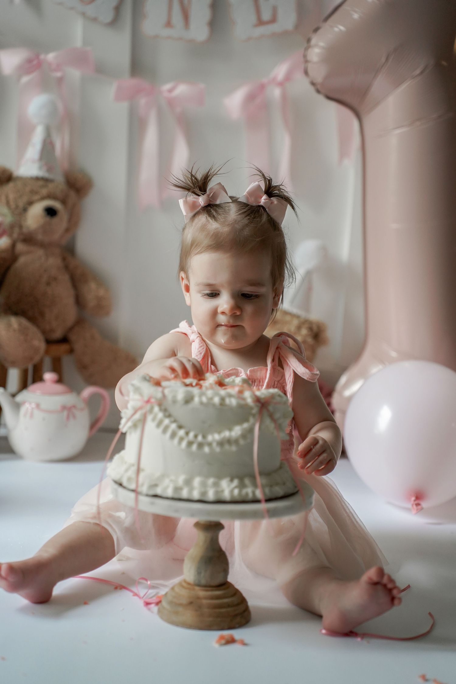 A pink and white themed cake smash photography session with a tutu dress, balloons, and teddy bear in the background.