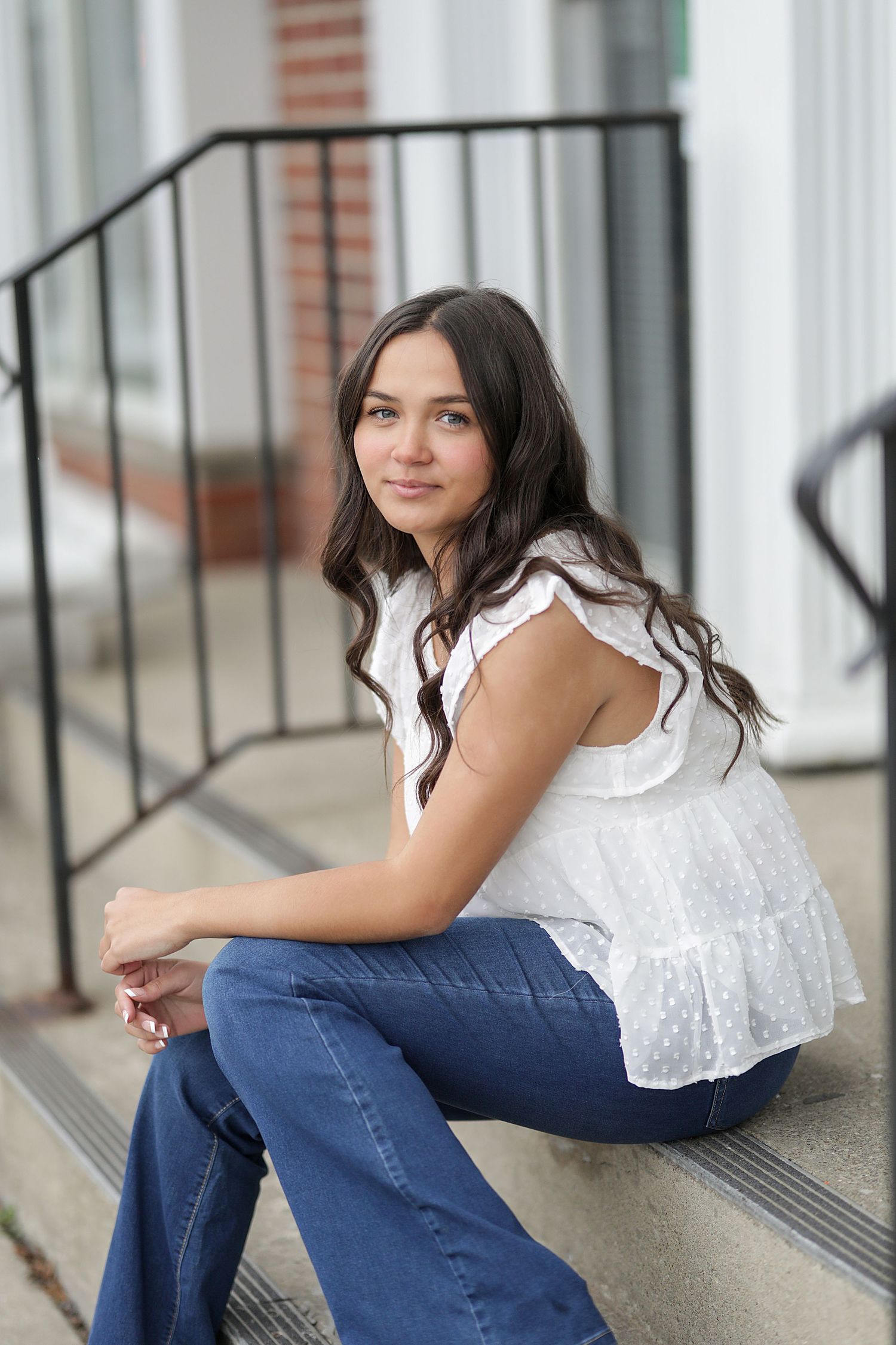 Young woman in a white lace top and blue jeans sits casually on outdoor concrete steps by a metal railing.
