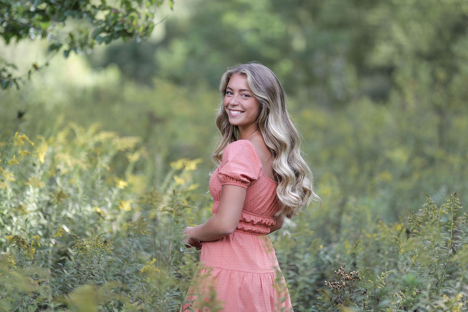 A woman wearing a coral pink dress twirls joyfully in a wildflower meadow during golden hour.