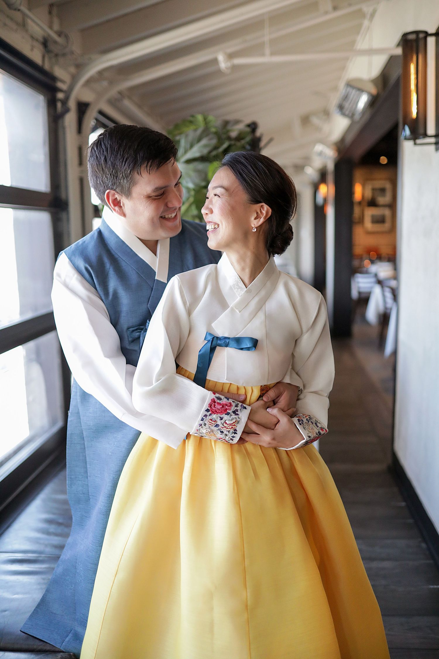 A couple embraces indoors wearing traditional wedding attire with a white hanbok and blue vest against a window-lit corridor.