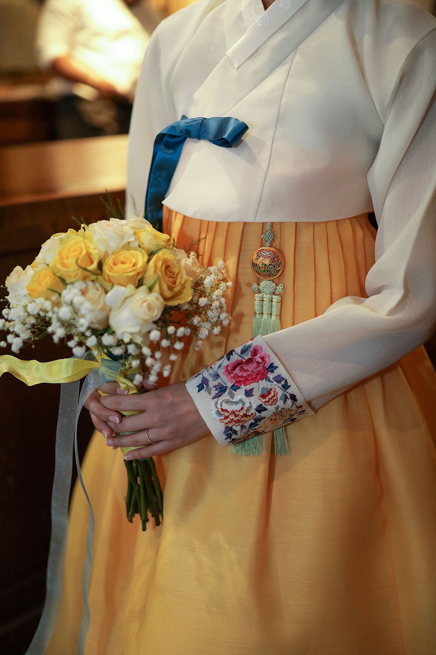 A person wearing a traditional Korean hanbok dress in yellow holds a bouquet of yellow roses and baby's breath.