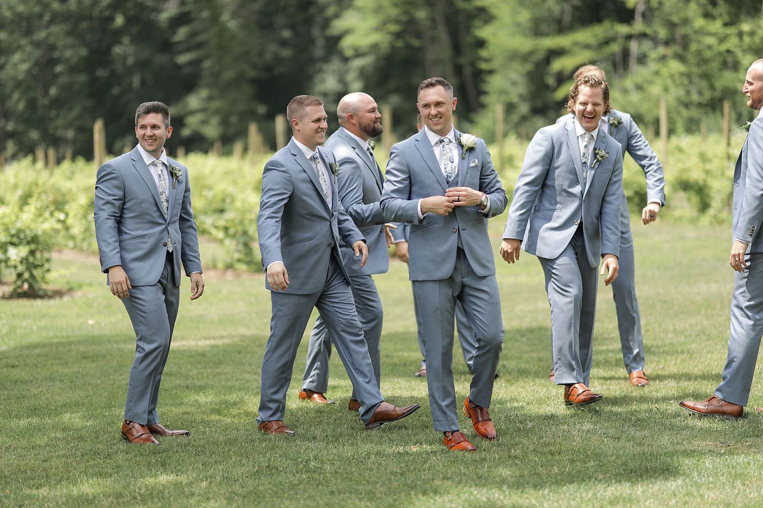 Wedding party members in grey suits enjoy a candid moment on manicured grounds.