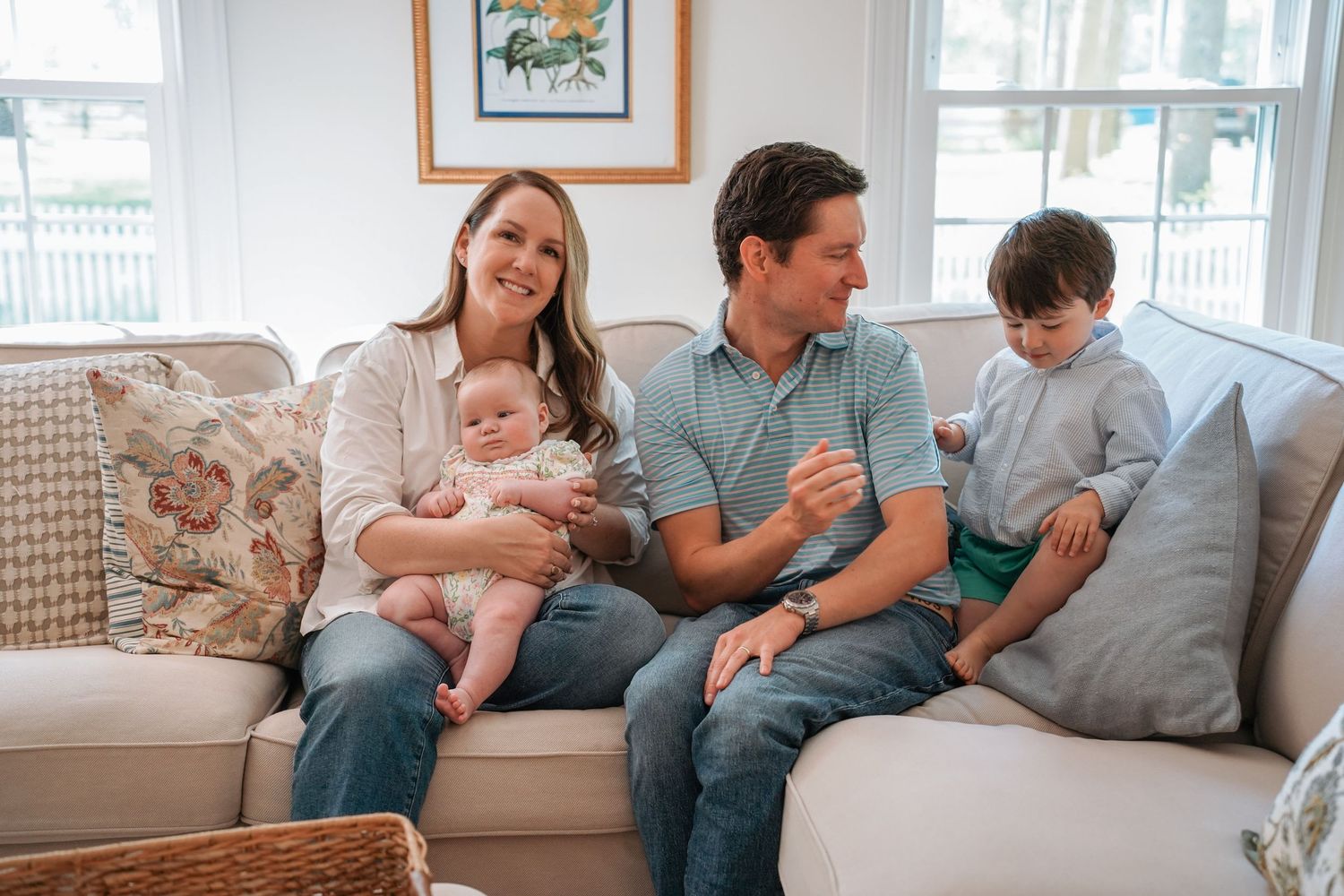 Family relaxes together on a gray couch in living room while sharing tender moments with small children.