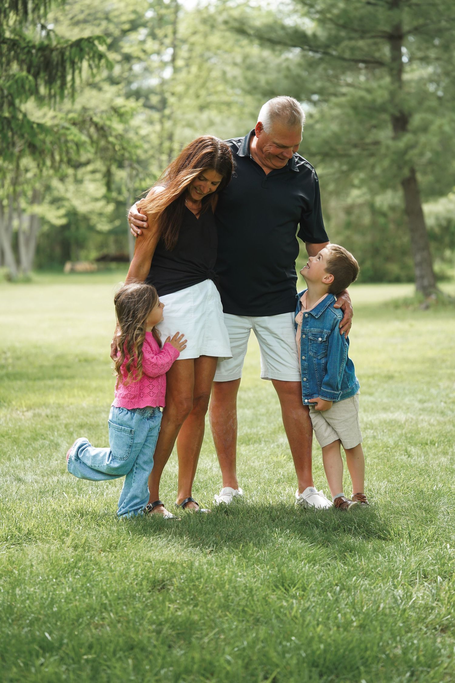A family shares an intimate moment together while standing on grass in a park with evergreen trees in the background.