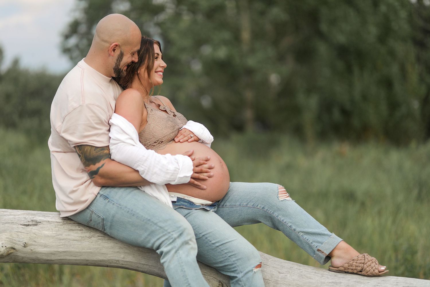 Expectant couple embraces while sitting on wooden bench in outdoor field setting during sunset. Maternity photographer Medina Ohio.