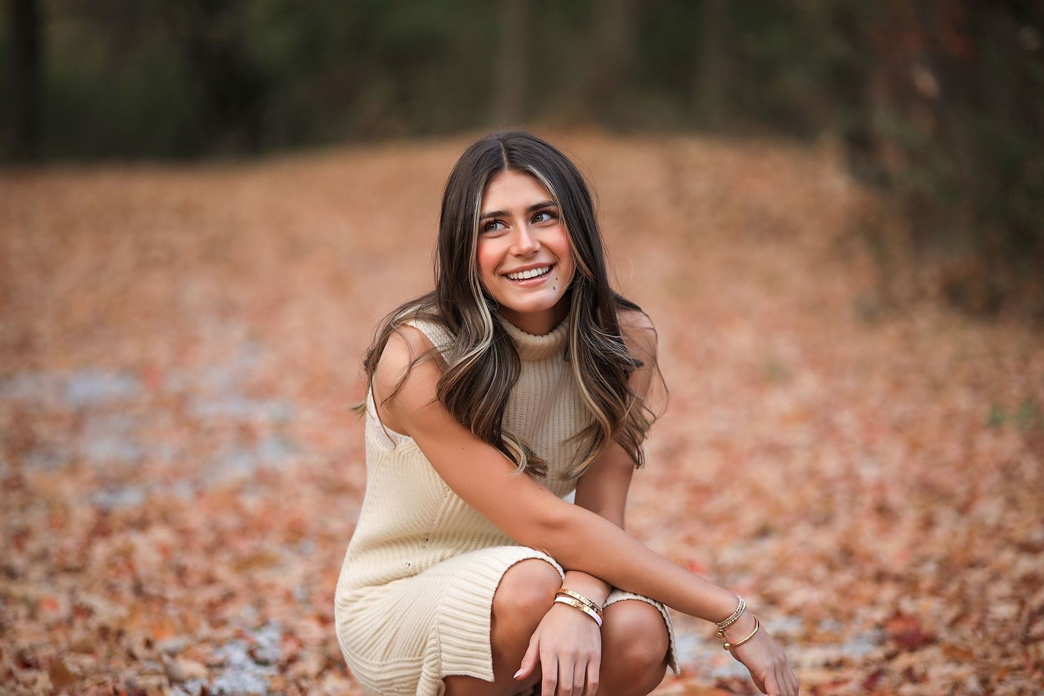 Person in cream colored dress sits on ground surrounded by fallen autumn leaves on forest floor and smiles warmly.