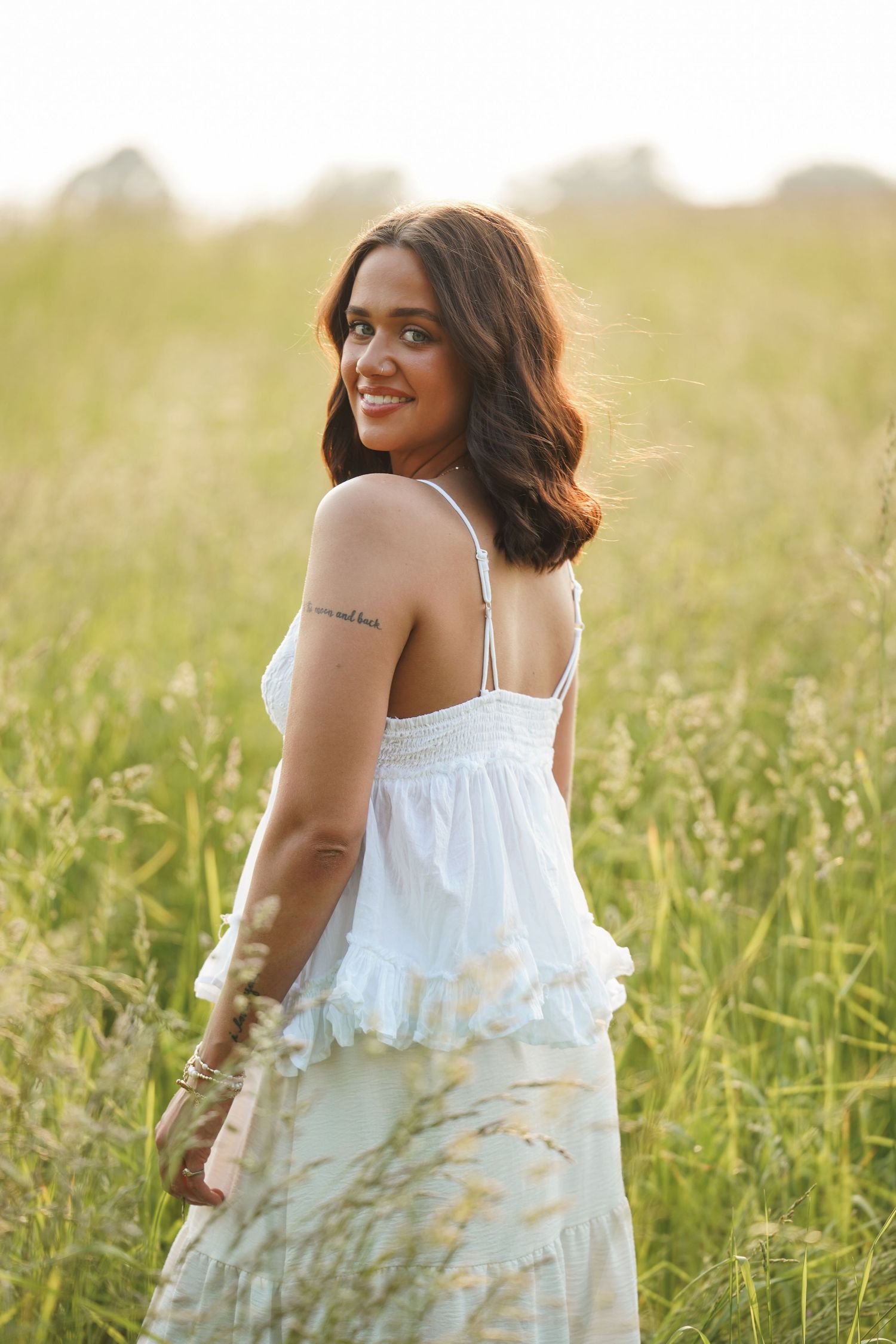 A woman in a flowing white sundress poses gracefully in a sunlit field of tall grass during golden hour.